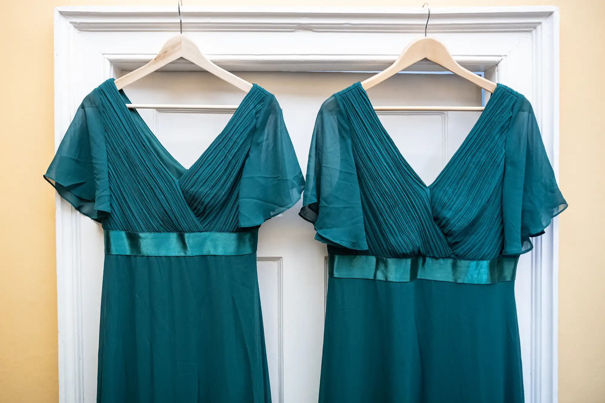 Two identical teal dresses with pleated V-neck bodices and flutter sleeves are hanging on hangers against a white door.