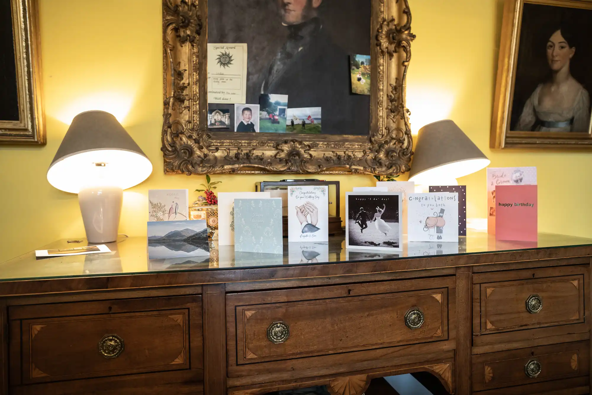 A wooden sideboard adorned with eight greeting cards and twin table lamps, set against a yellow wall with framed portrait paintings.