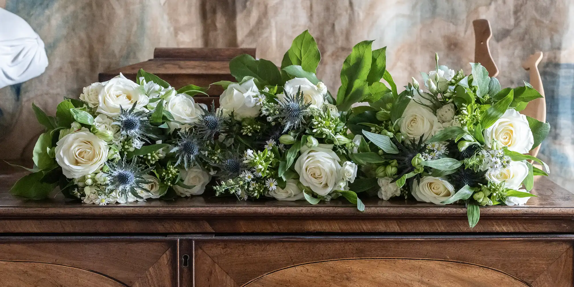 Three floral arrangements featuring white roses, green foliage, and thistle flowers placed on a wooden cabinet.