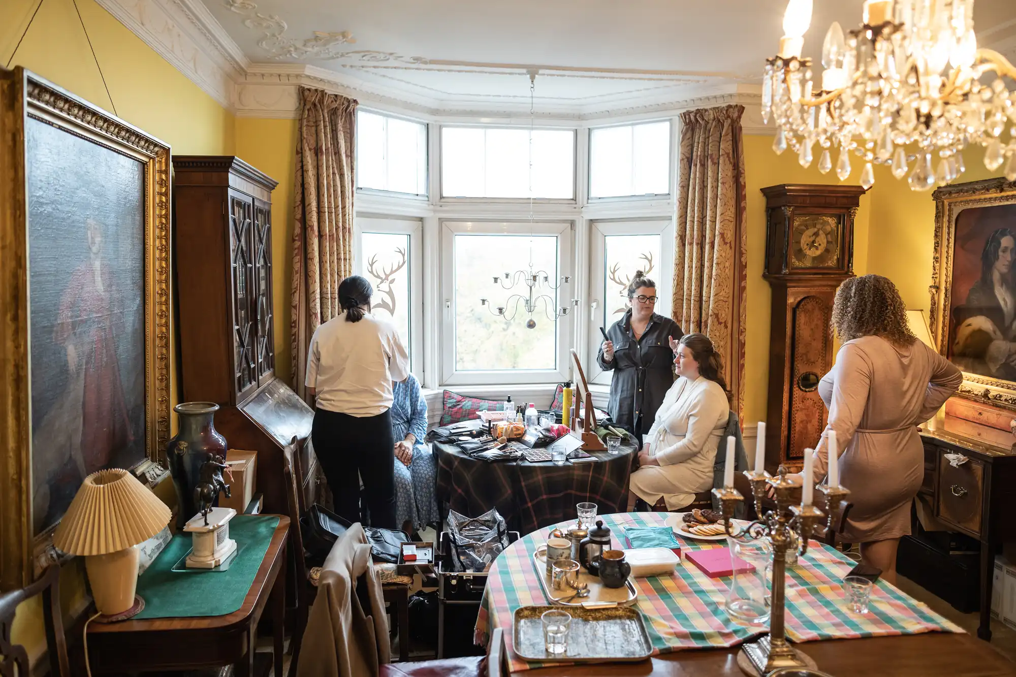 People in a vintage-decorated room, gathered around a table with various items. Two individuals appear to be preparing costumes with antlers for an upcoming event or production.