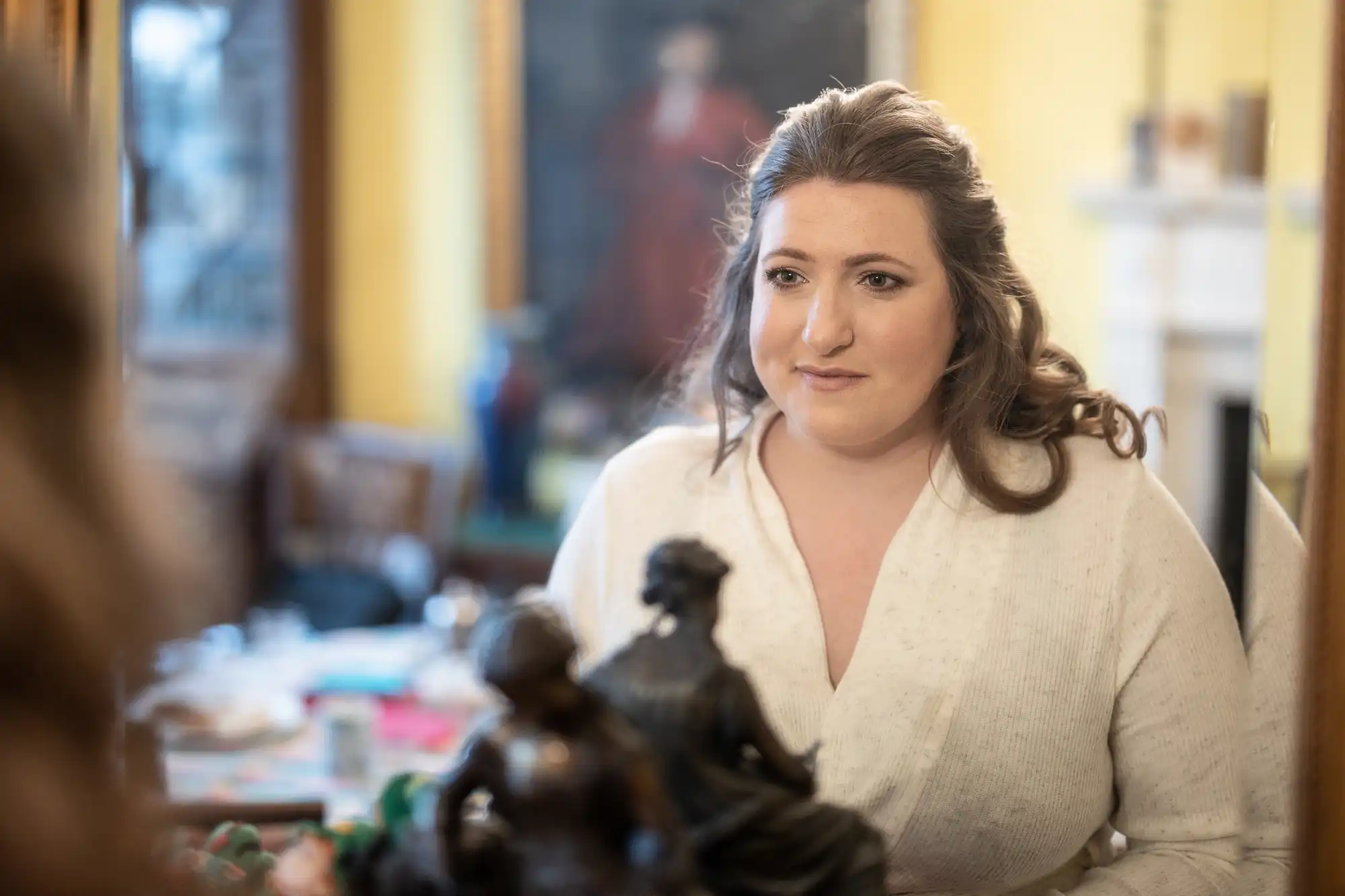 A woman with long hair and a white top looks at herself in a mirror in a softly lit room with blurred furniture and a statue in the foreground.