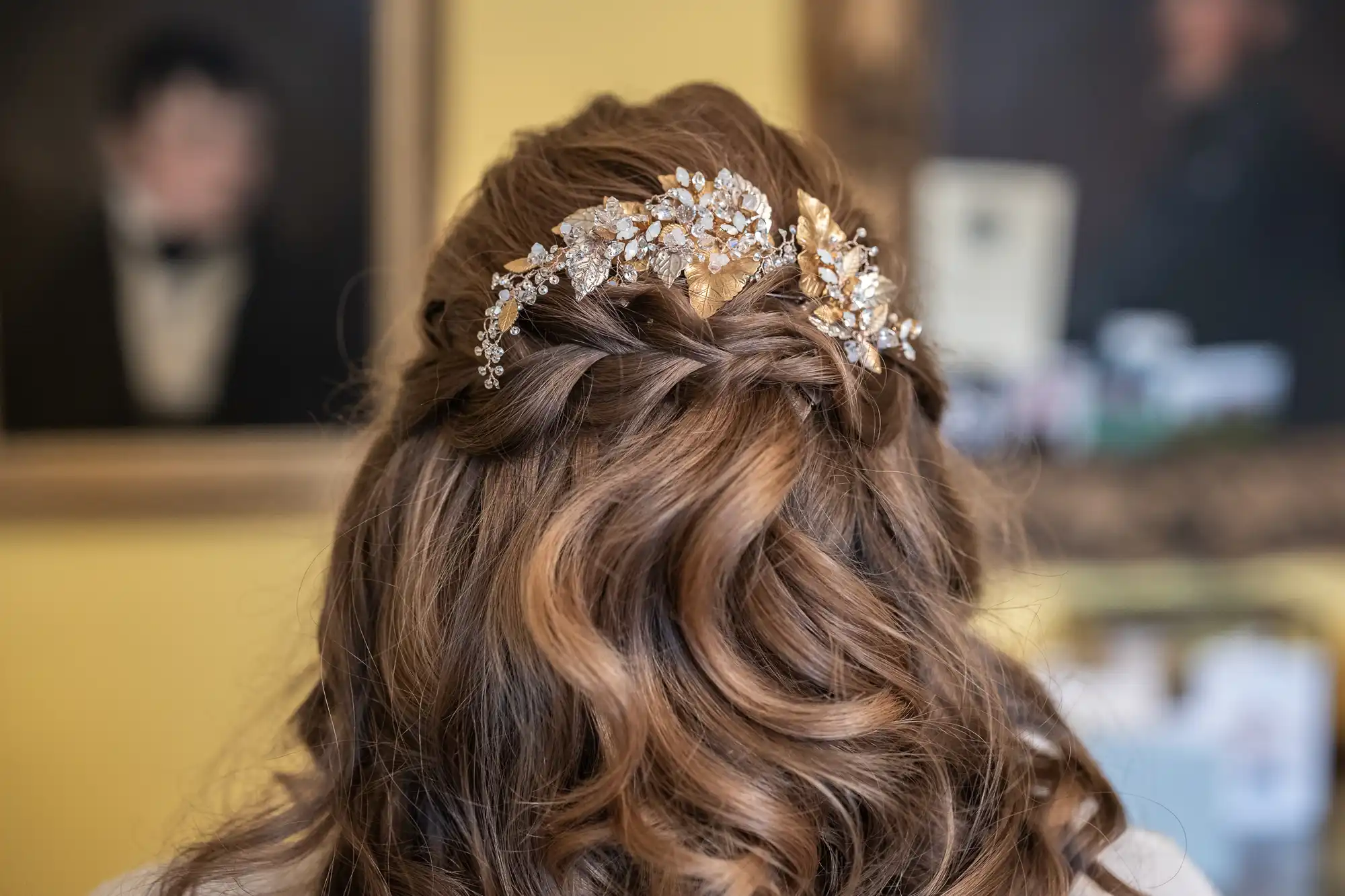 A woman with wavy brown hair is seen from the back, wearing a decorative gold and silver floral hairpiece with intricate details. Blurred portraits are visible in the background.