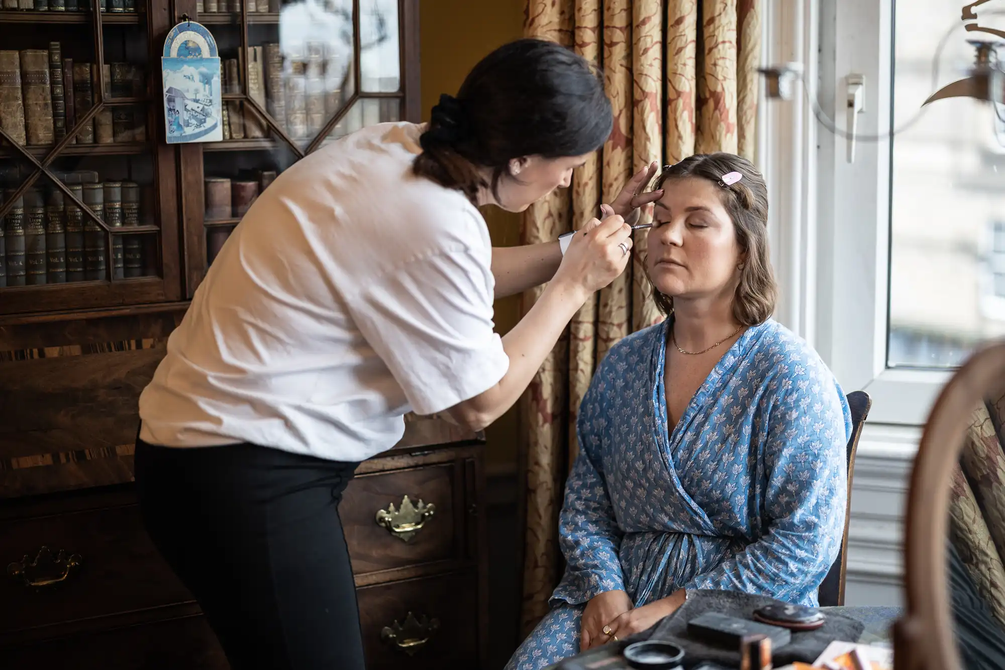 A woman in a white shirt applies makeup to another woman seated in a floral robe, in a room with wooden furniture and a window with curtains.