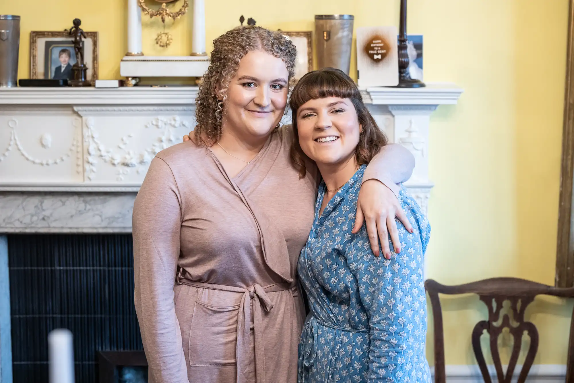 Two people standing close together, smiling, with one arm around the other. They are indoors in front of a decorated mantlepiece with framed photos and other items.