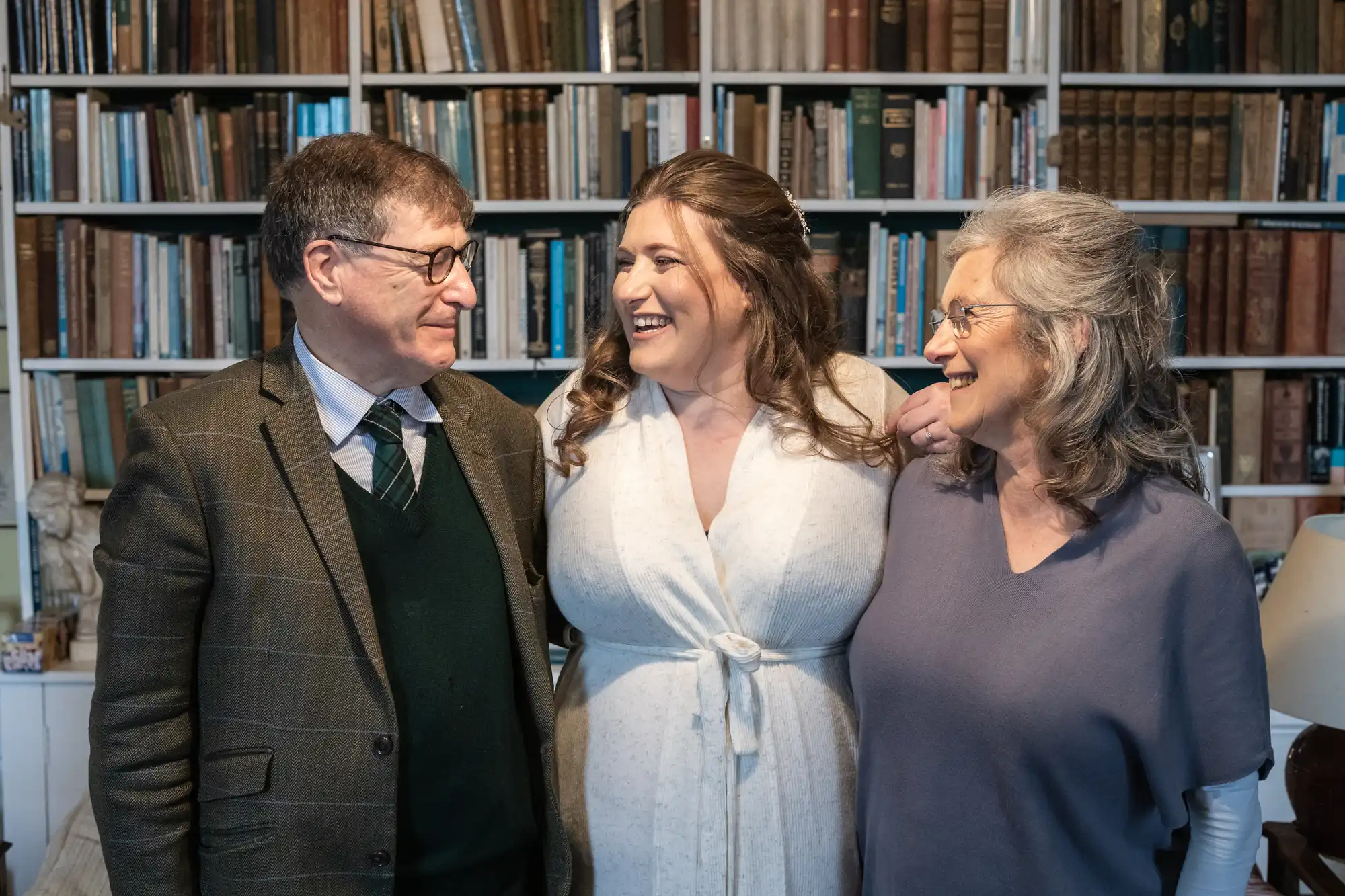 Three people stand closely together in front of a bookshelf. They are smiling and appear to be engaged in conversation. The person in the center is wearing a white dress.