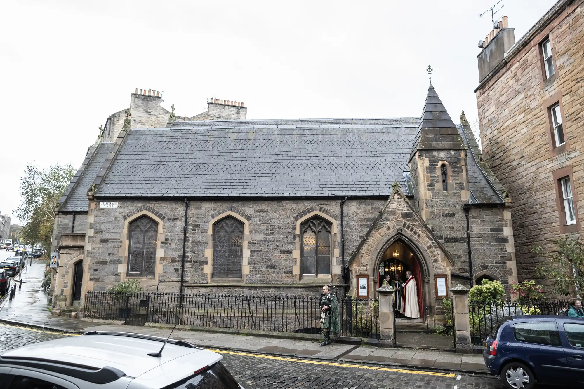 A stone church with a grey slate roof stands at a street corner. Two people are under an arch by the entrance. The scene includes a parked blue car and other buildings in the background.