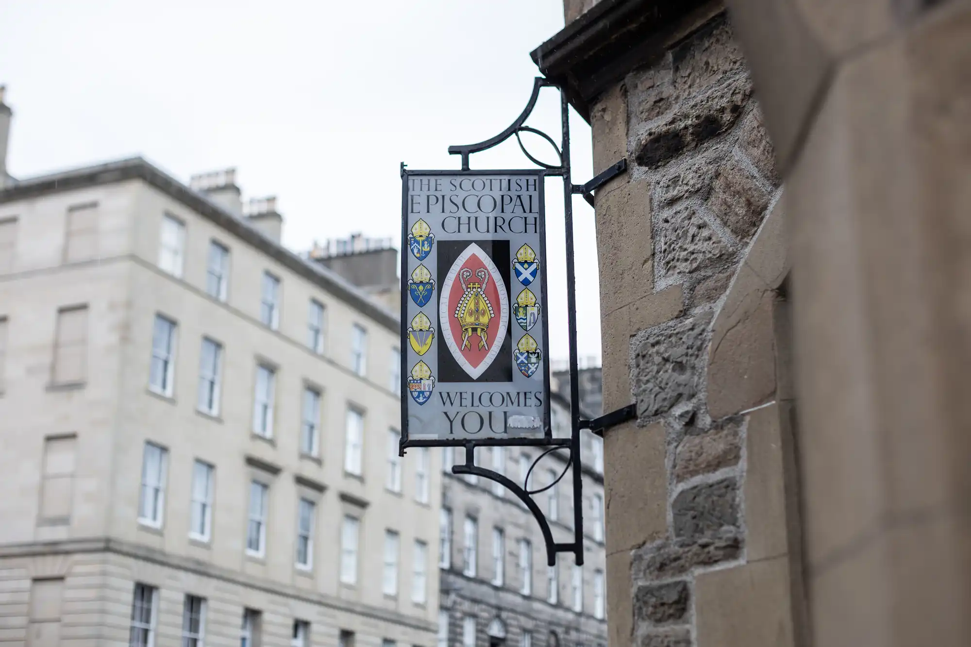 A sign attached to a stone building reads, "The Scottish Episcopal Church welcomes you," with a crest in the center. Nearby buildings with numerous windows are visible in the background.