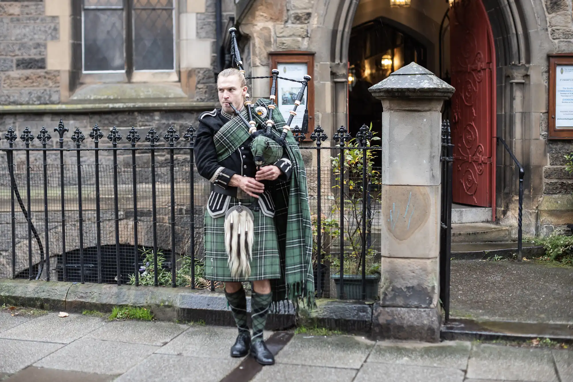 A man in traditional Scottish attire plays the bagpipes on a sidewalk next to a stone building with a red door and metal railing.