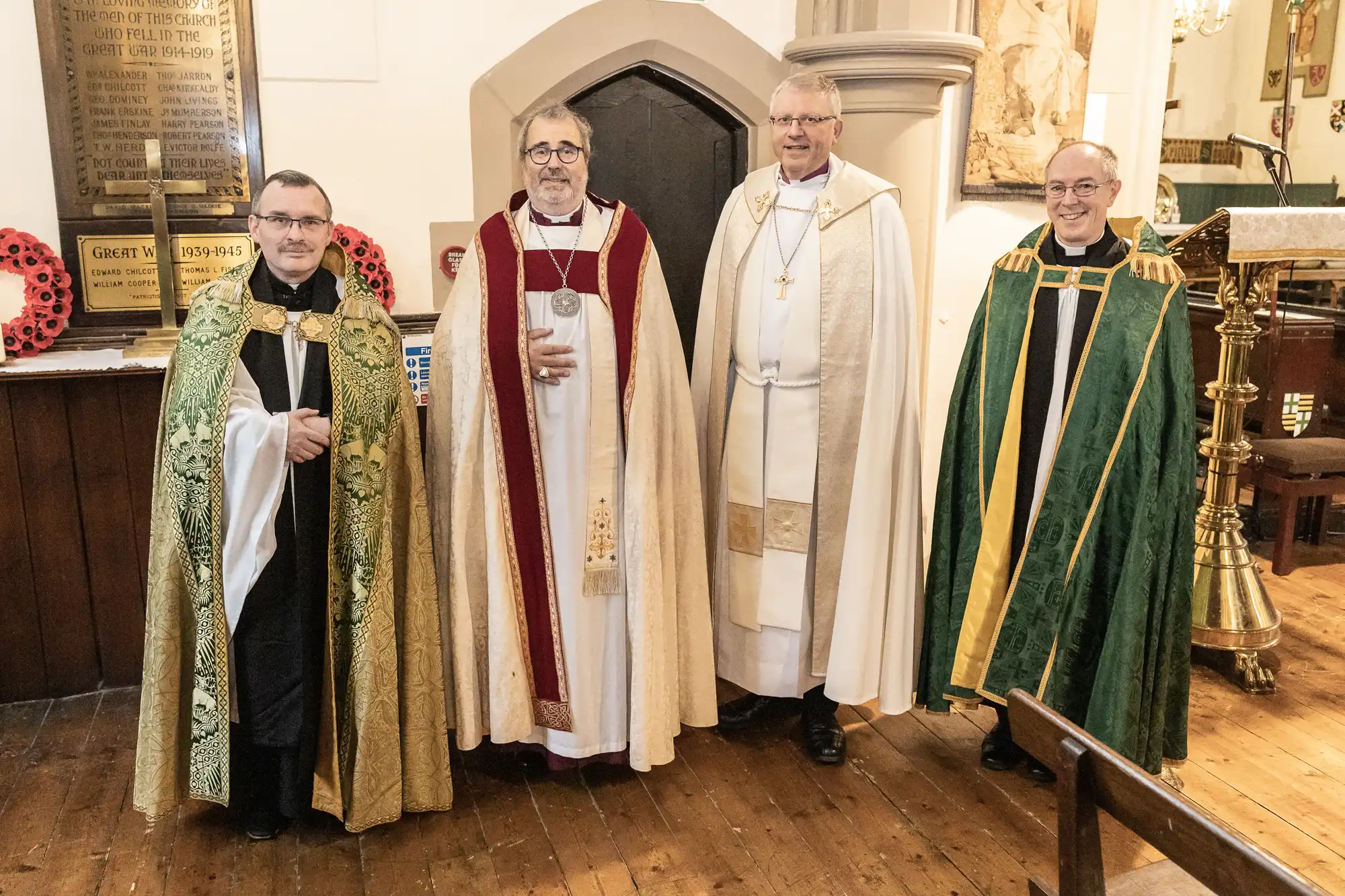 Four men dressed in religious vestments stand together in a church. They wear robes of green, red, white, and green, respectively, with various symbols and decorations.