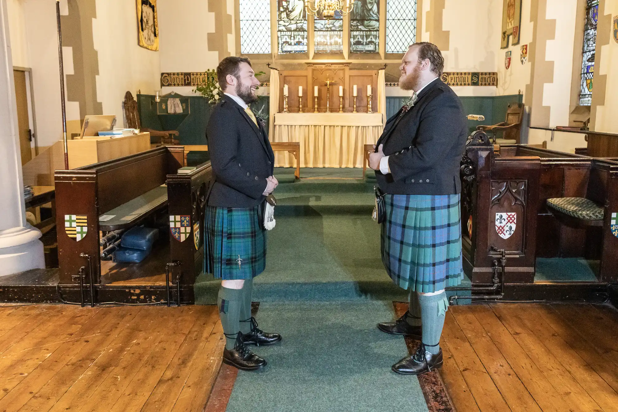 Two men in traditional Scottish attire stand facing each other inside a church with wooden floors and a central aisle leading to the altar.