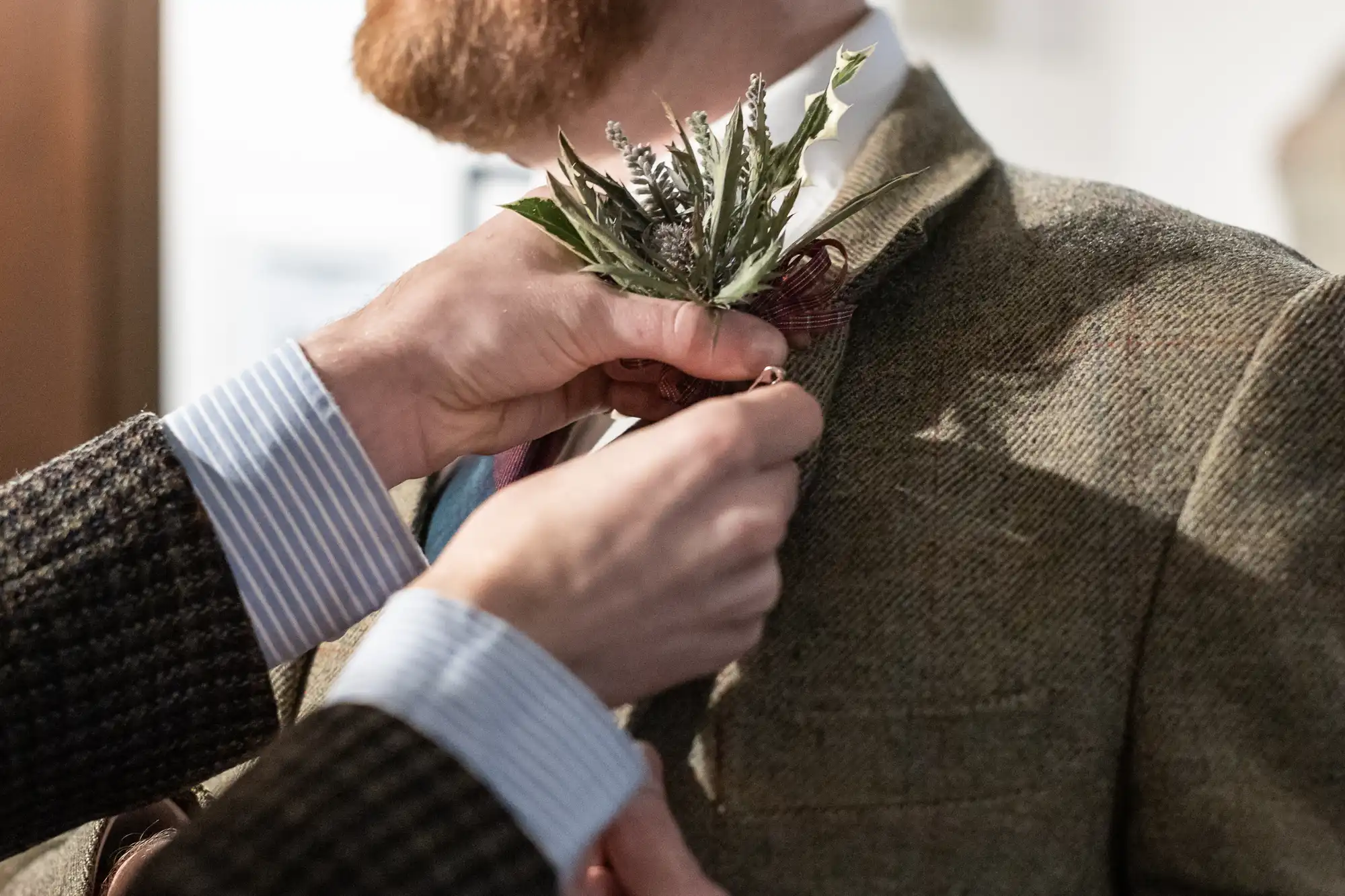 A person secures a floral boutonnière to the lapel of a man wearing a textured suit jacket.