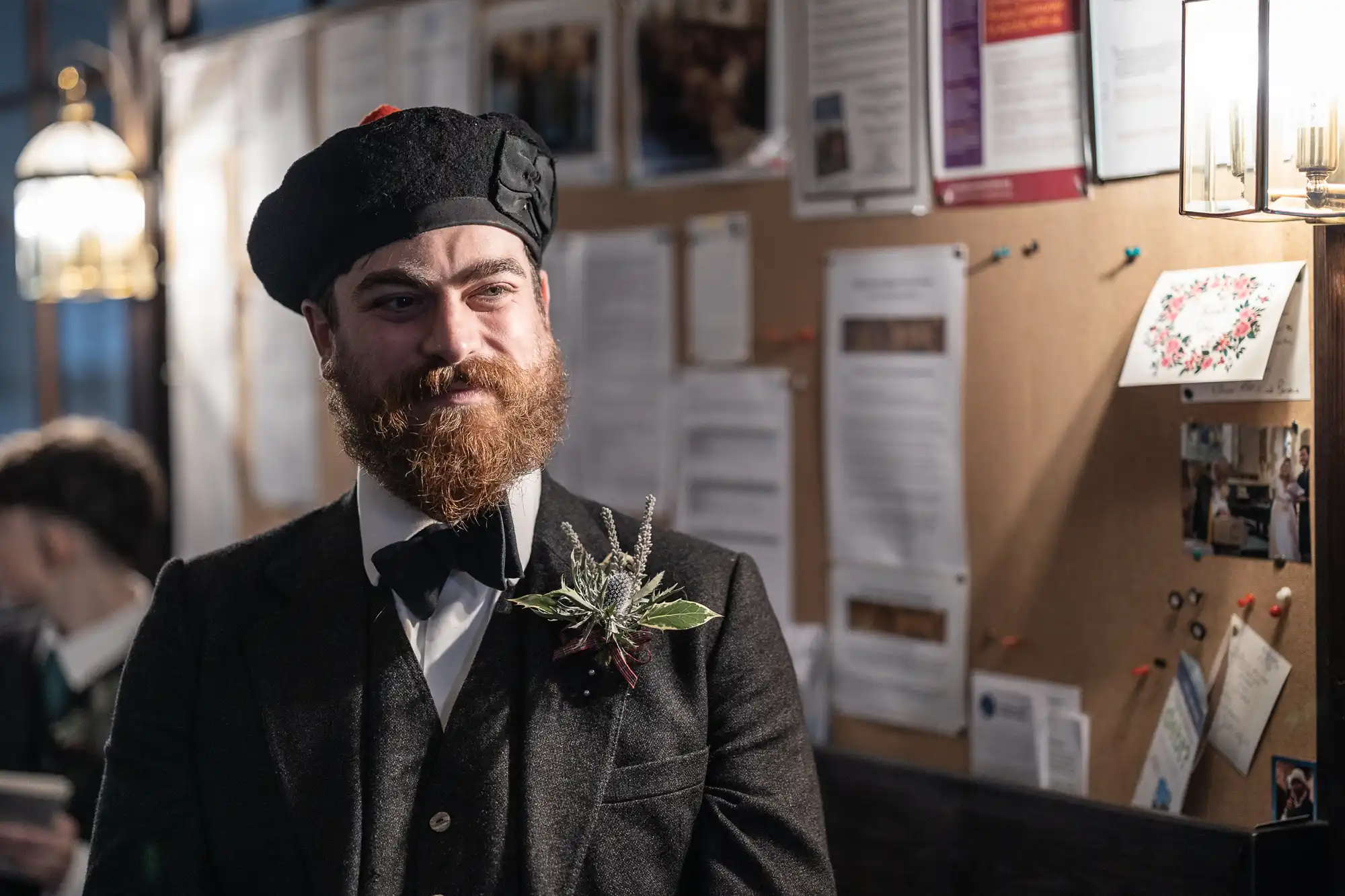 A bearded man wearing a dark beret, suit, and bowtie stands indoors in front of a bulletin board covered with papers and notes.