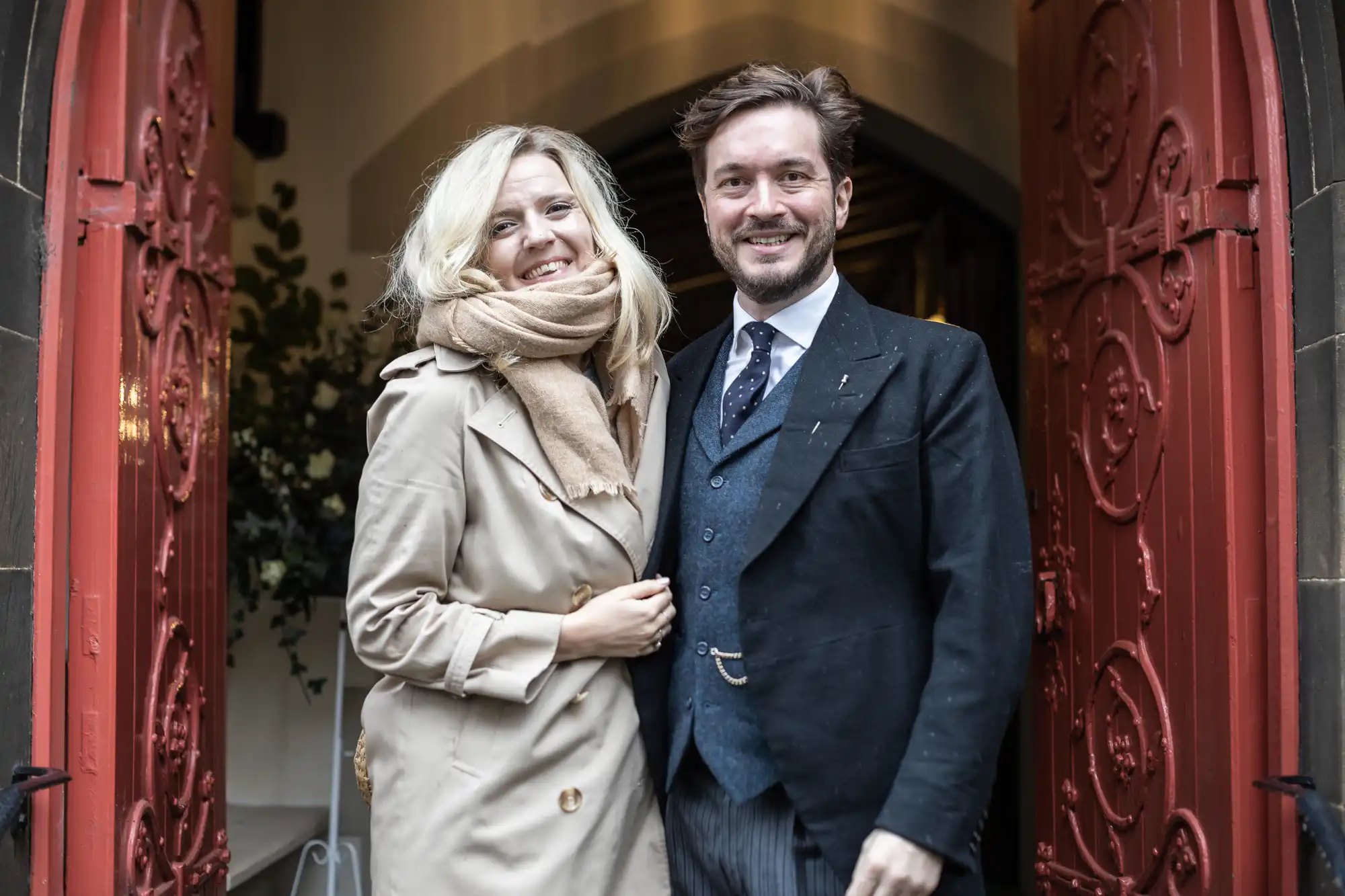 A man and a woman stand together, smiling, in the doorway of a building with ornate red doors. The man is wearing a suit and the woman is wearing a beige coat and scarf.