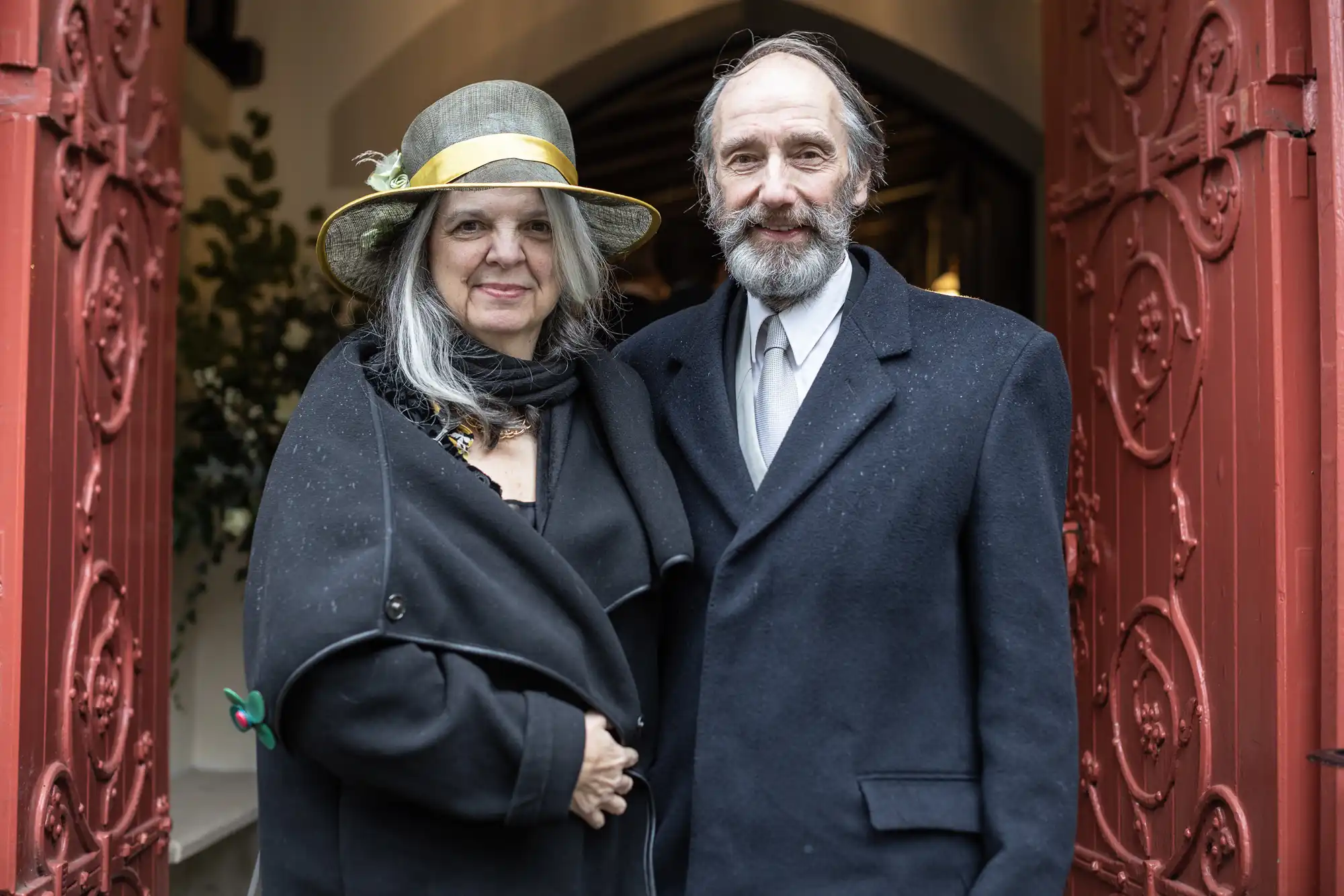 An elderly couple poses together in front of a building with open red doors; the woman is wearing a hat and dark coat, while the man wears a suit and tie.