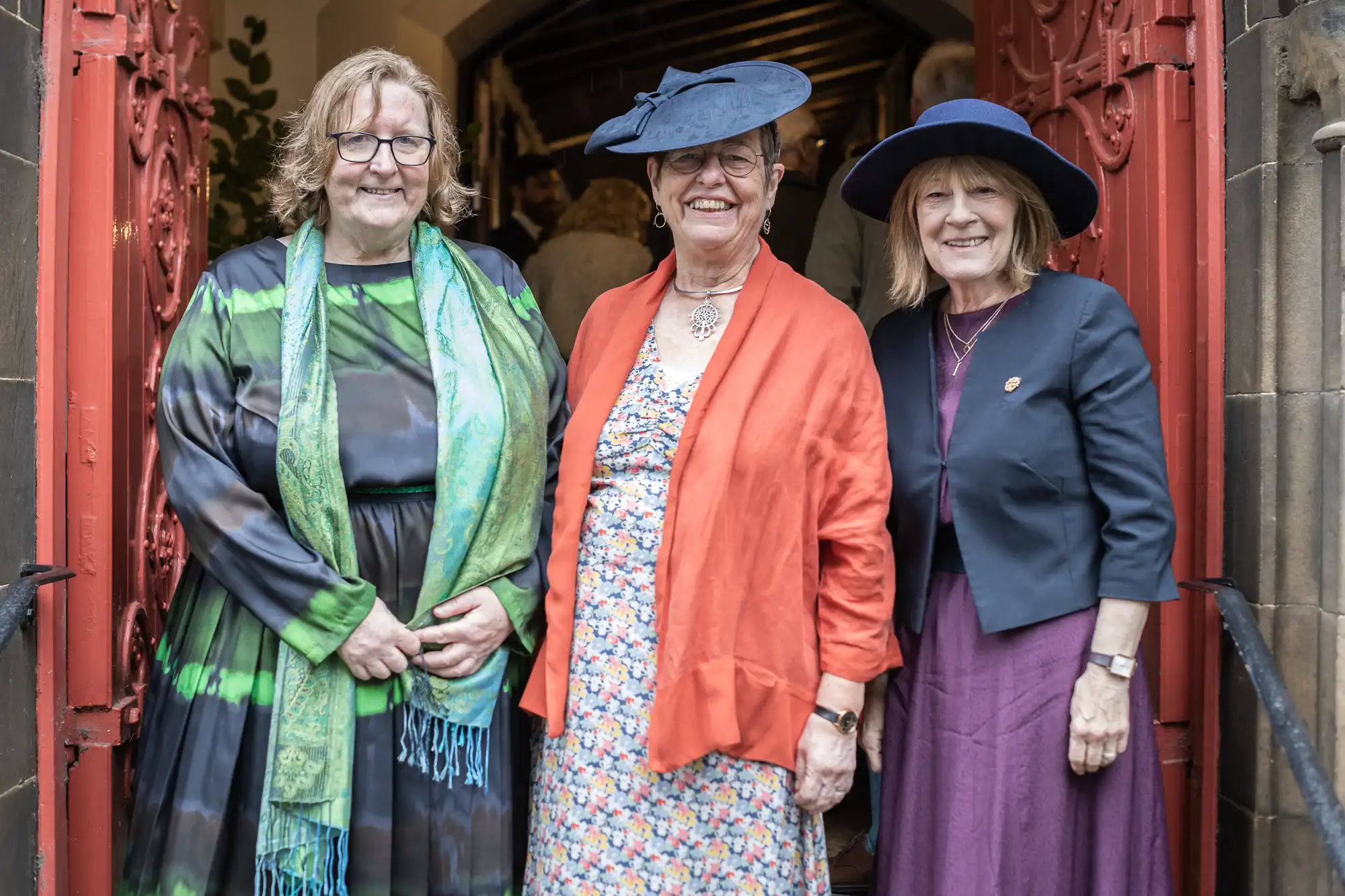 Three women dressed in formal attire with hats stand in front of a red door, smiling at the camera.