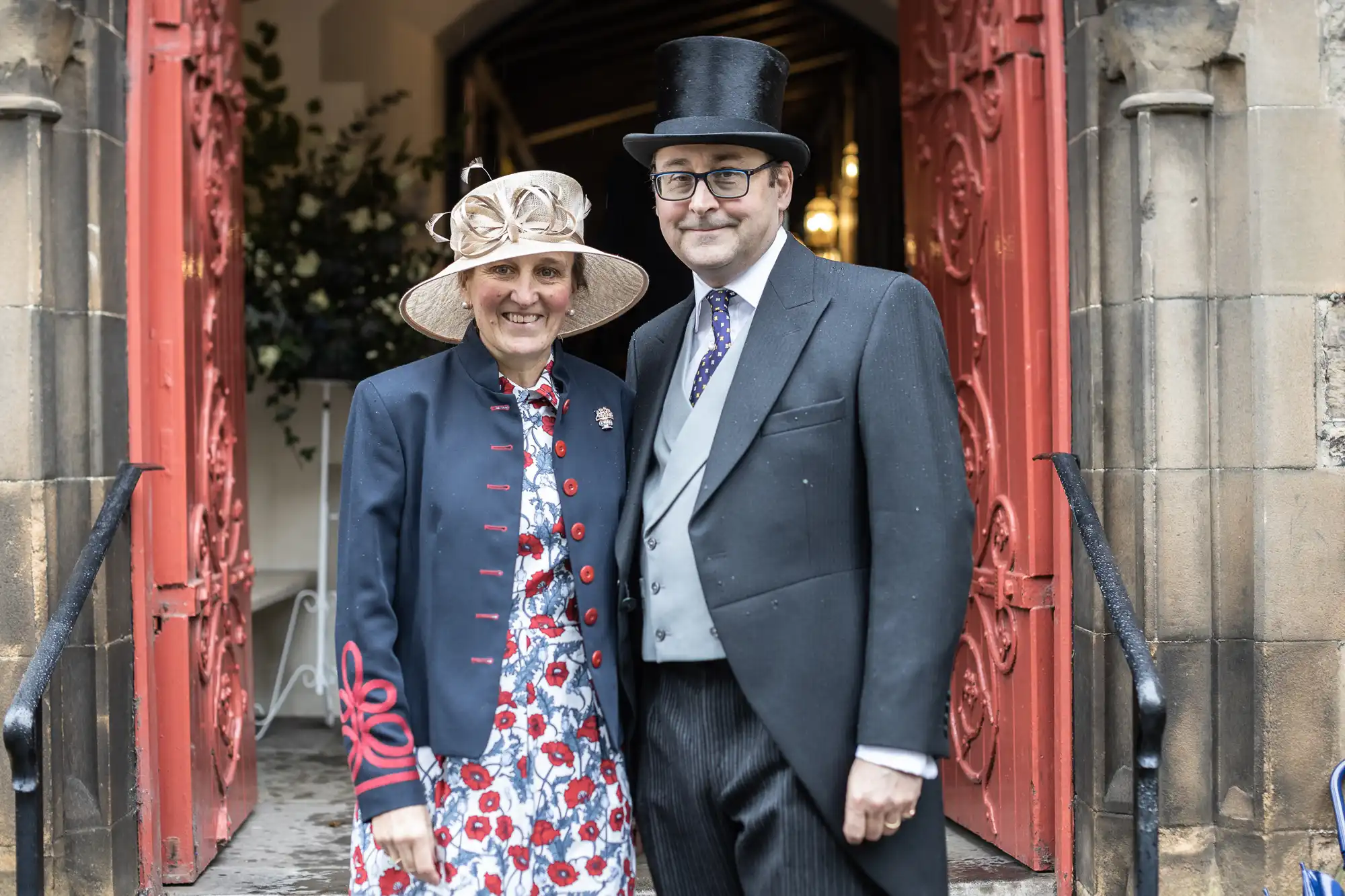 Two individuals dressed in formal attire, including hats, standing side by side in front of a red, open, arched doorway.