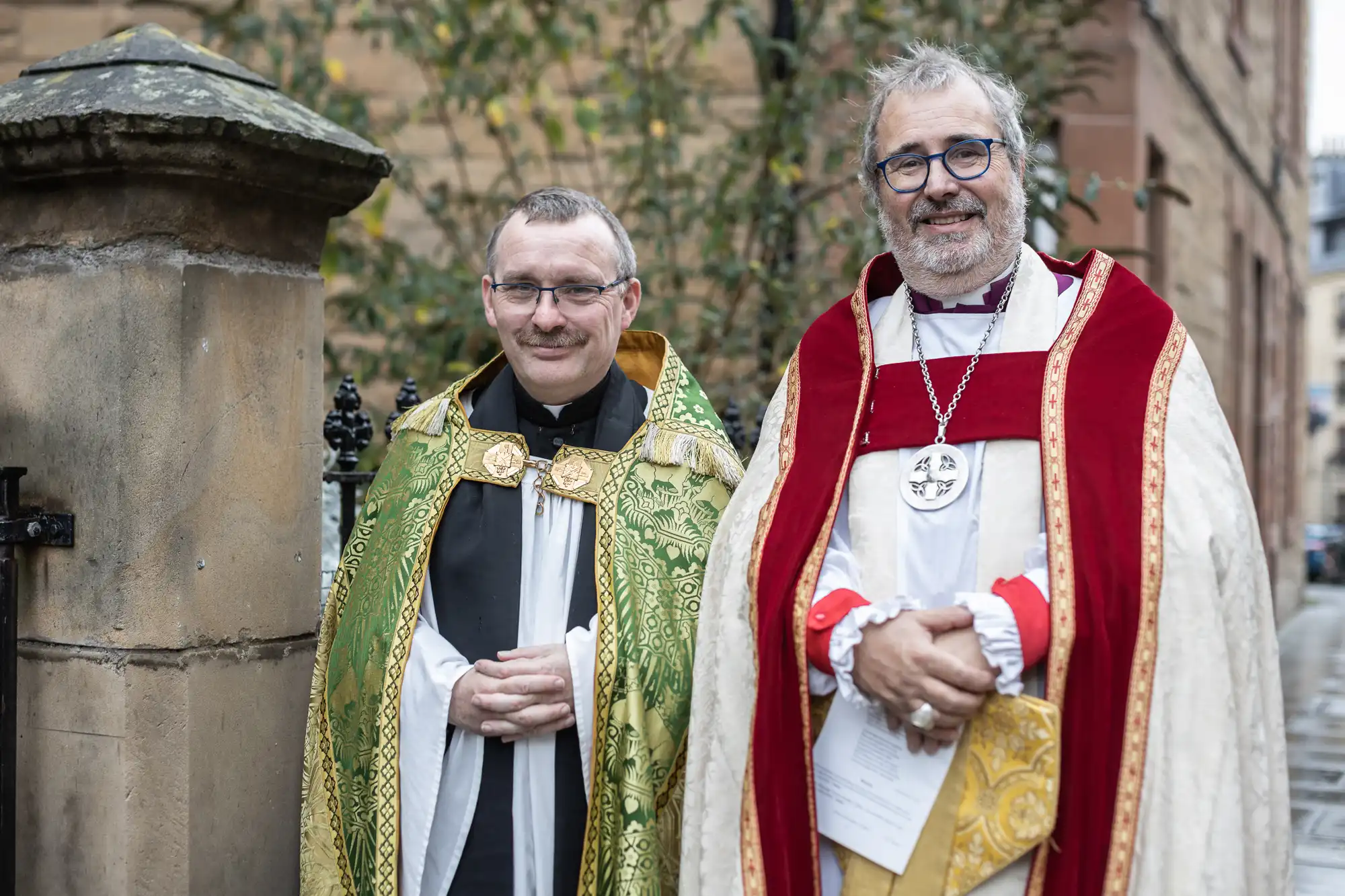 Two clergy members in ceremonial robes stand outdoors, smiling at the camera. One wears green and gold attire, the other red and white. A stone wall and greenery are visible in the background.