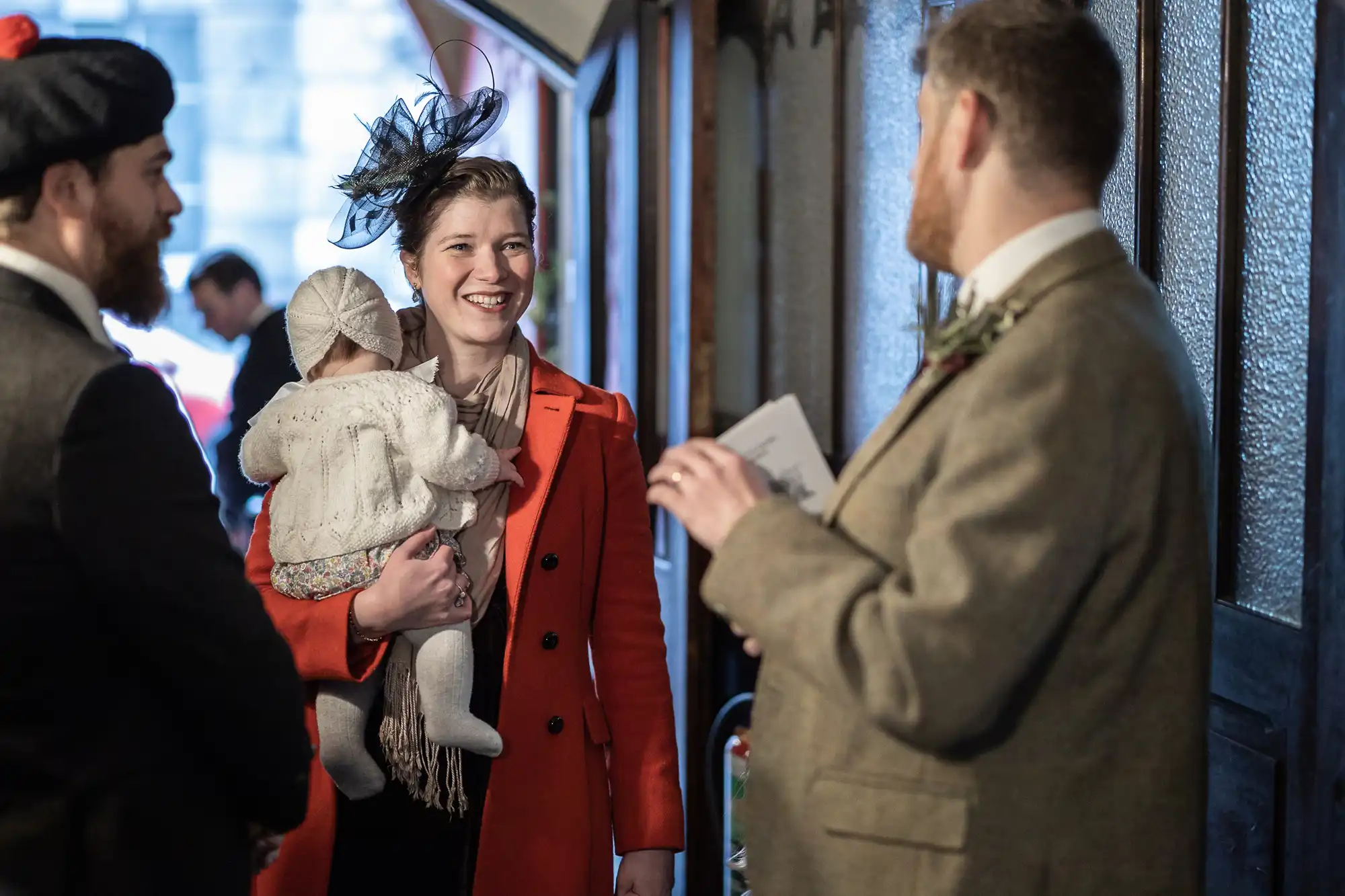 Three adults, one holding a baby in a white outfit, engage in conversation indoors. One adult is wearing a bright orange coat and a black hat with an adornment.