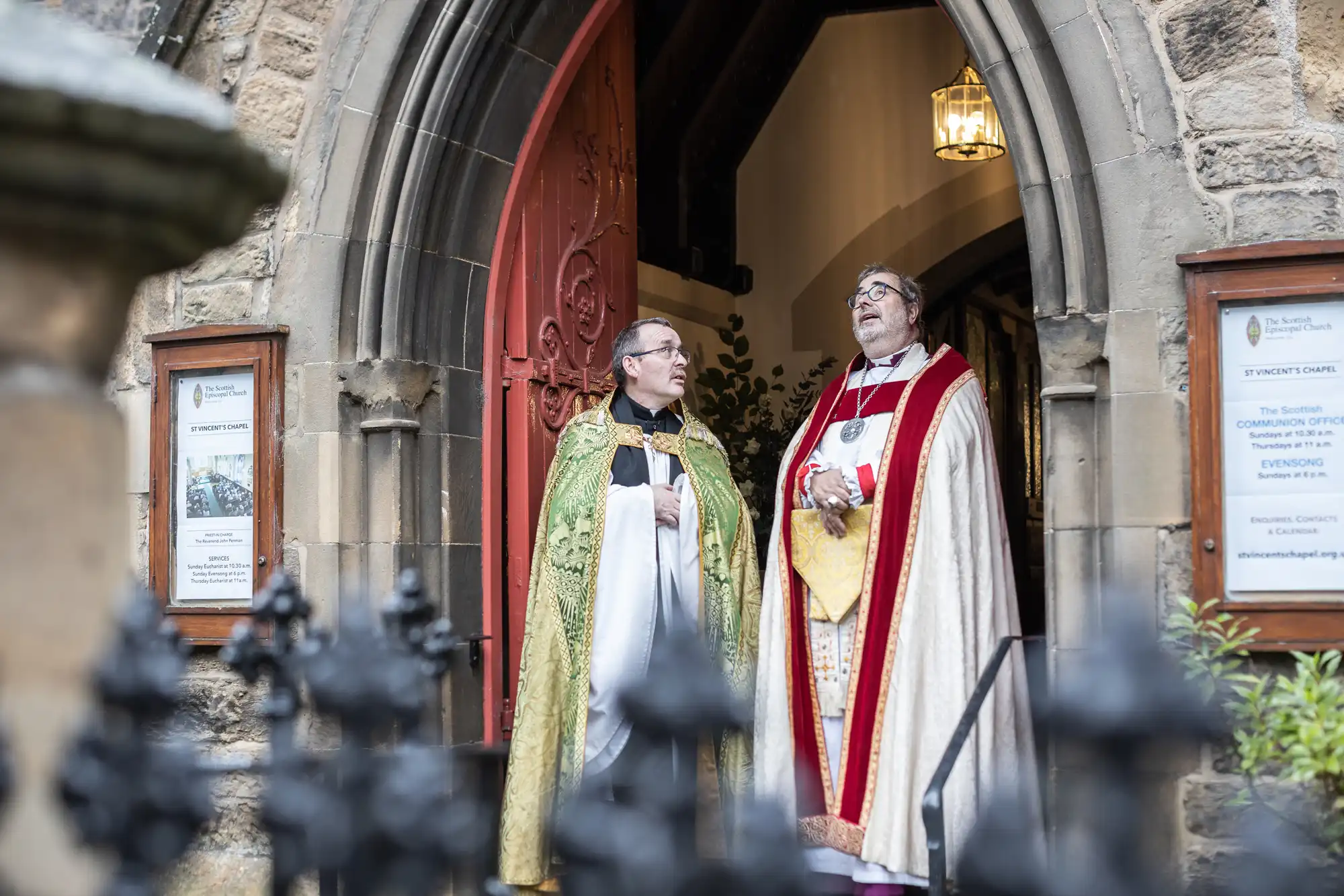 Two clergy members in ceremonial robes stand in the doorway of a stone church, engaged in conversation. The archway includes a red door, and there are notices and posters on either side.