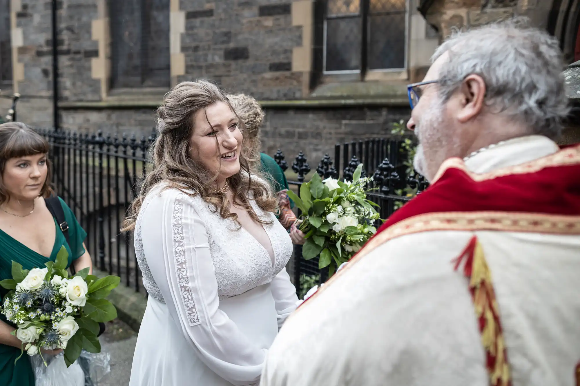 A bride in a white dress smiles while speaking with an elderly man in ceremonial attire outside a stone building. Another woman in a green dress stands nearby holding a bouquet.