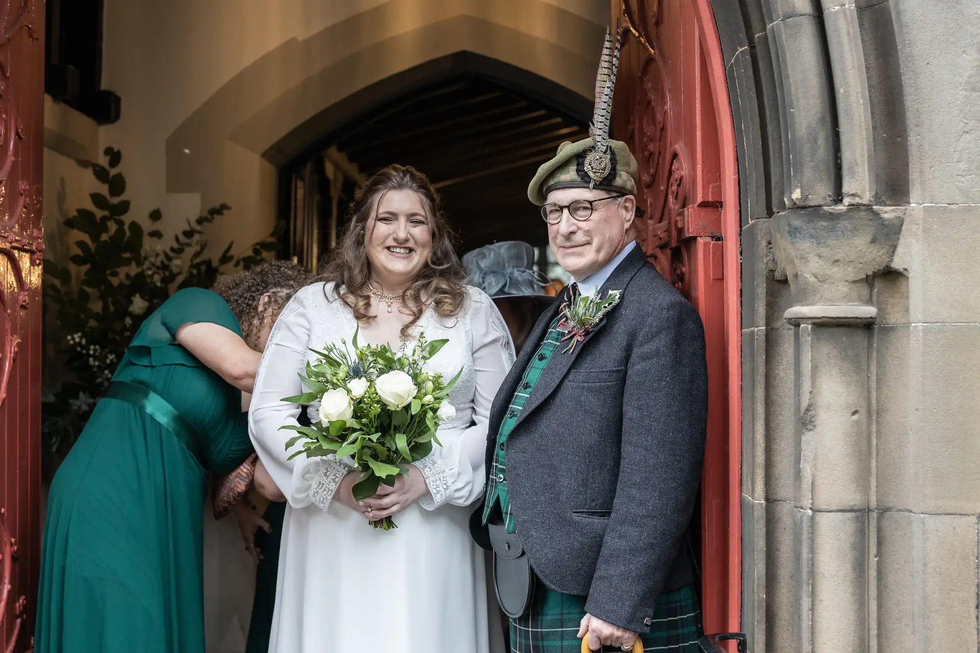 A bride and an older man are standing at the entrance of a building. The bride holds a bouquet of flowers and wears a white dress, while the man wears traditional Scottish attire, including a kilt and a hat.