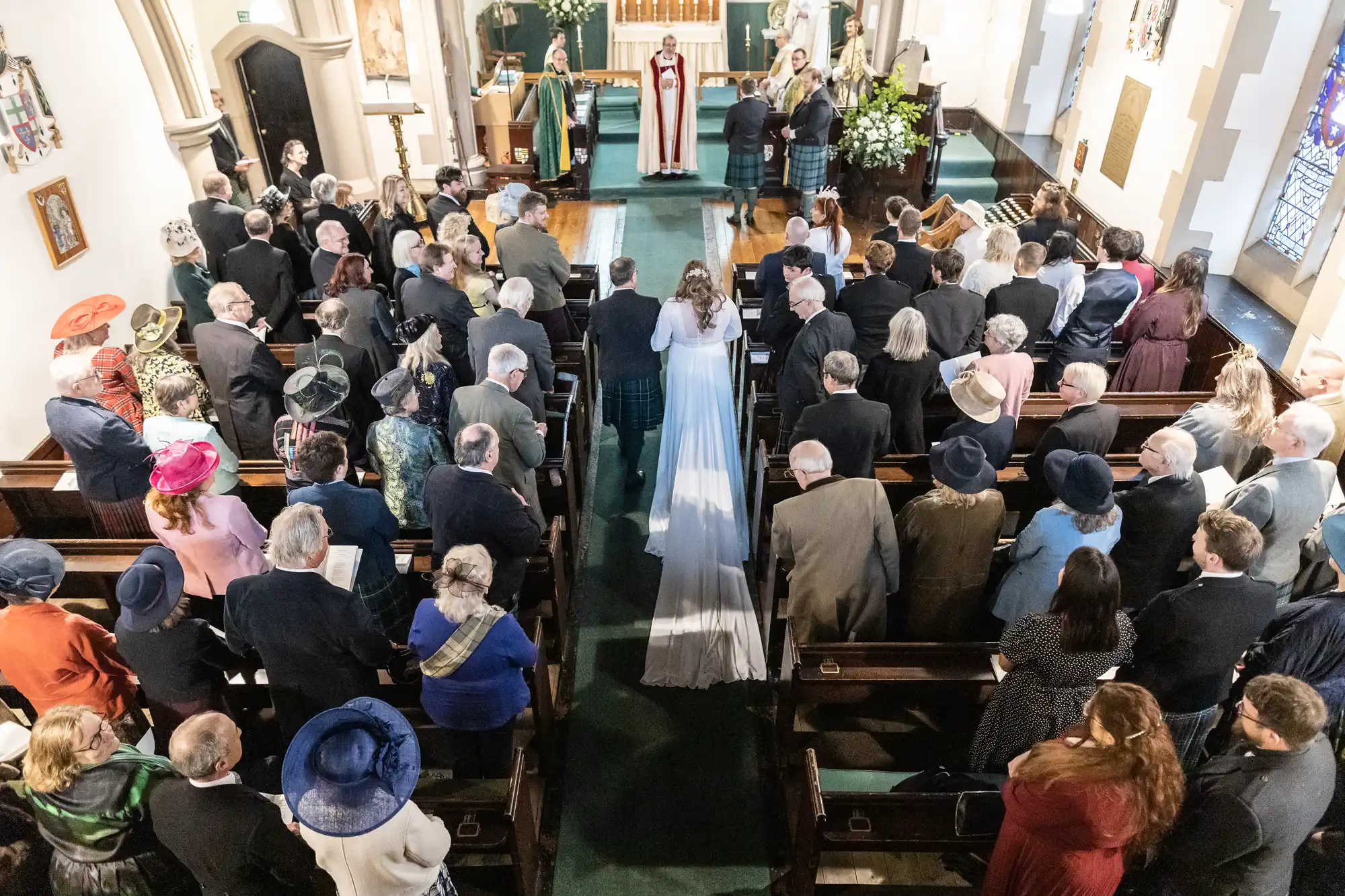 A bride in a white gown walks down the aisle of a church, accompanied by a man. Many guests are seated and standing, dressed formally. Clergy members are visible at the altar.