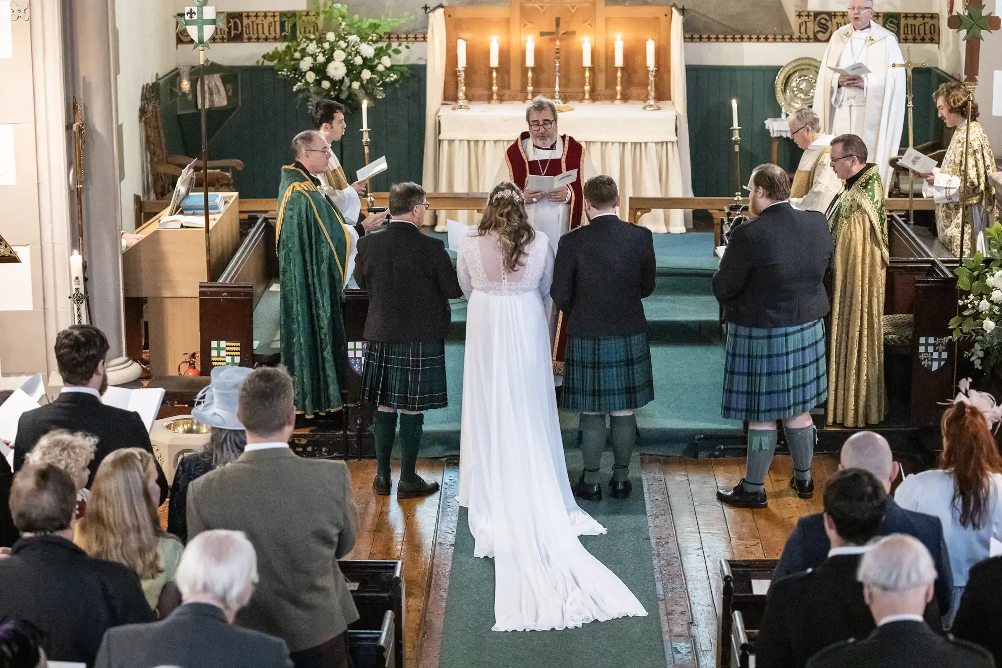 A couple stands before a priest during a wedding ceremony in a church. The groom and groomsmen wear kilts, while the bride wears a white gown. Clergy members are in the background. People are seated facing them.