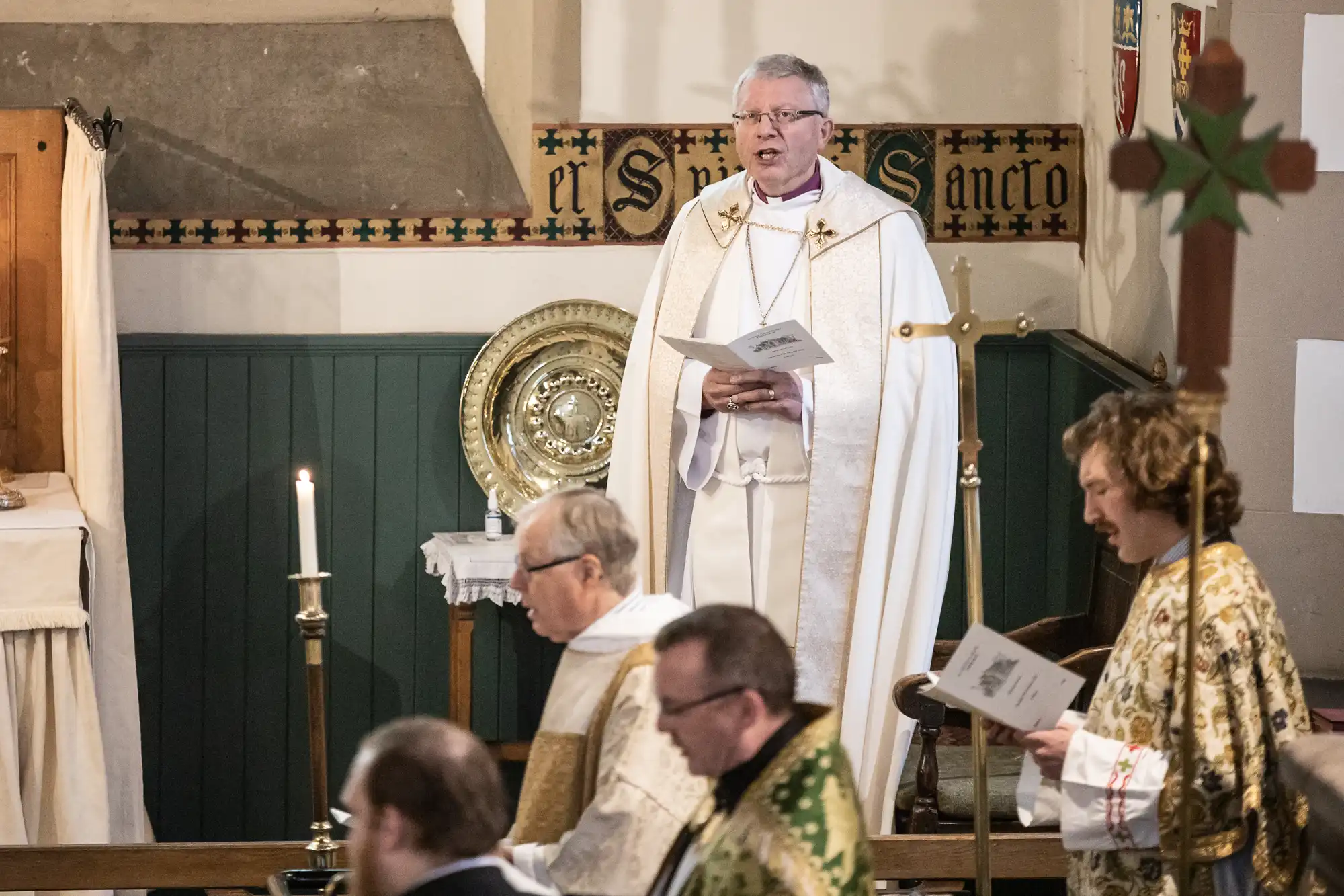 A clergyman in white robes speaks at a church ceremony. Other clergy members in various robes are seated and holding papers. Candles and religious symbols are visible in the background.