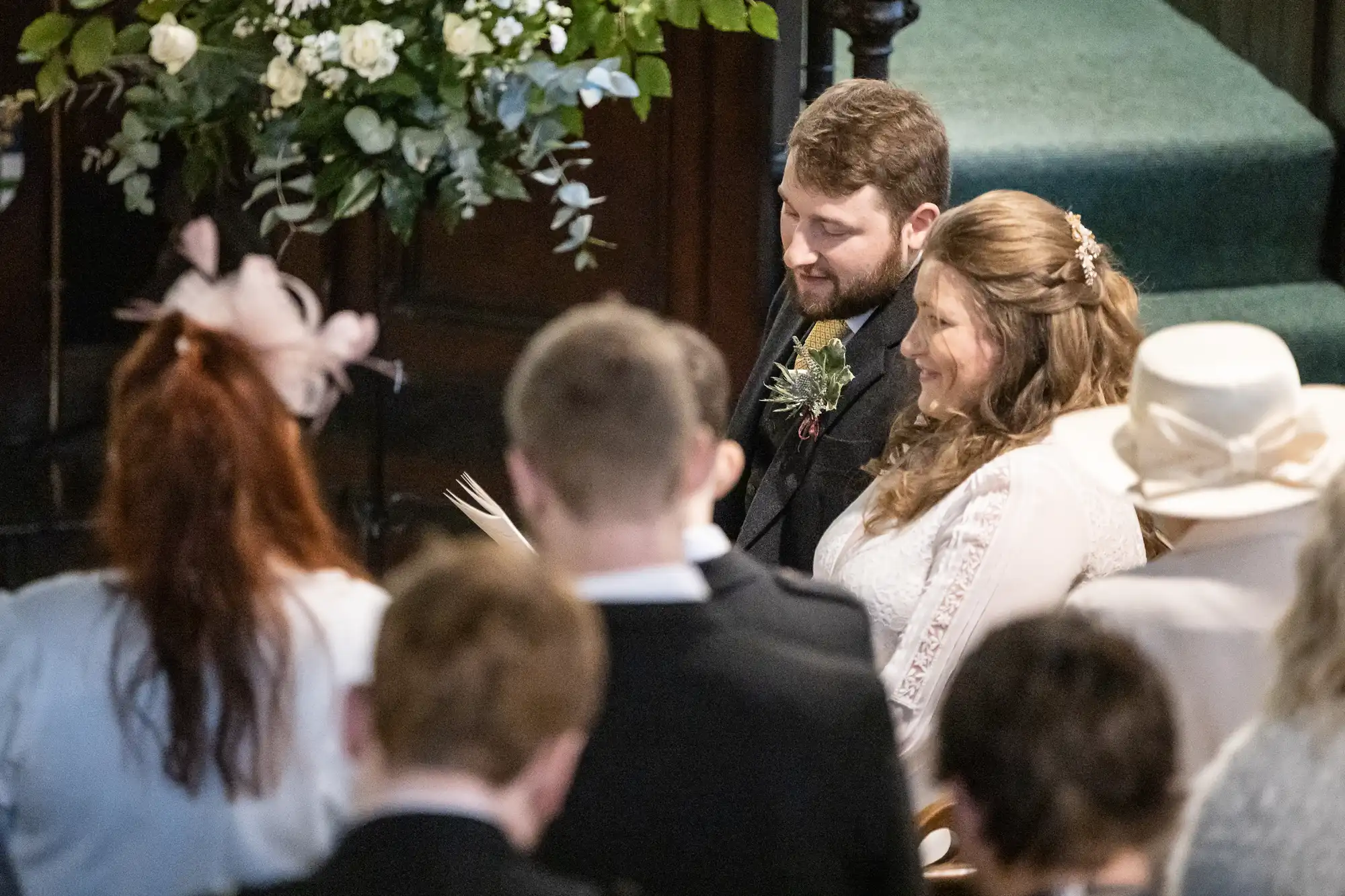 A bride and groom are seated during a wedding ceremony, surrounded by guests. The bride is wearing a white dress, and the groom is in a suit with a boutonniere.