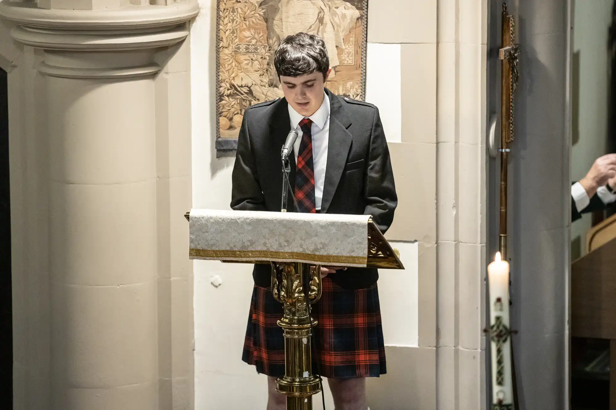 A person wearing a kilt and suit jacket is standing at a podium in a church, speaking into a microphone. A lit candle is visible in the foreground.