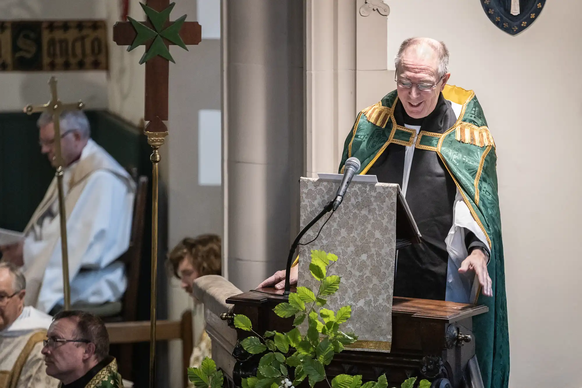 A clergyman in green and gold vestments speaks at a podium in a church. Other clergy members in white robes are seated in the background.