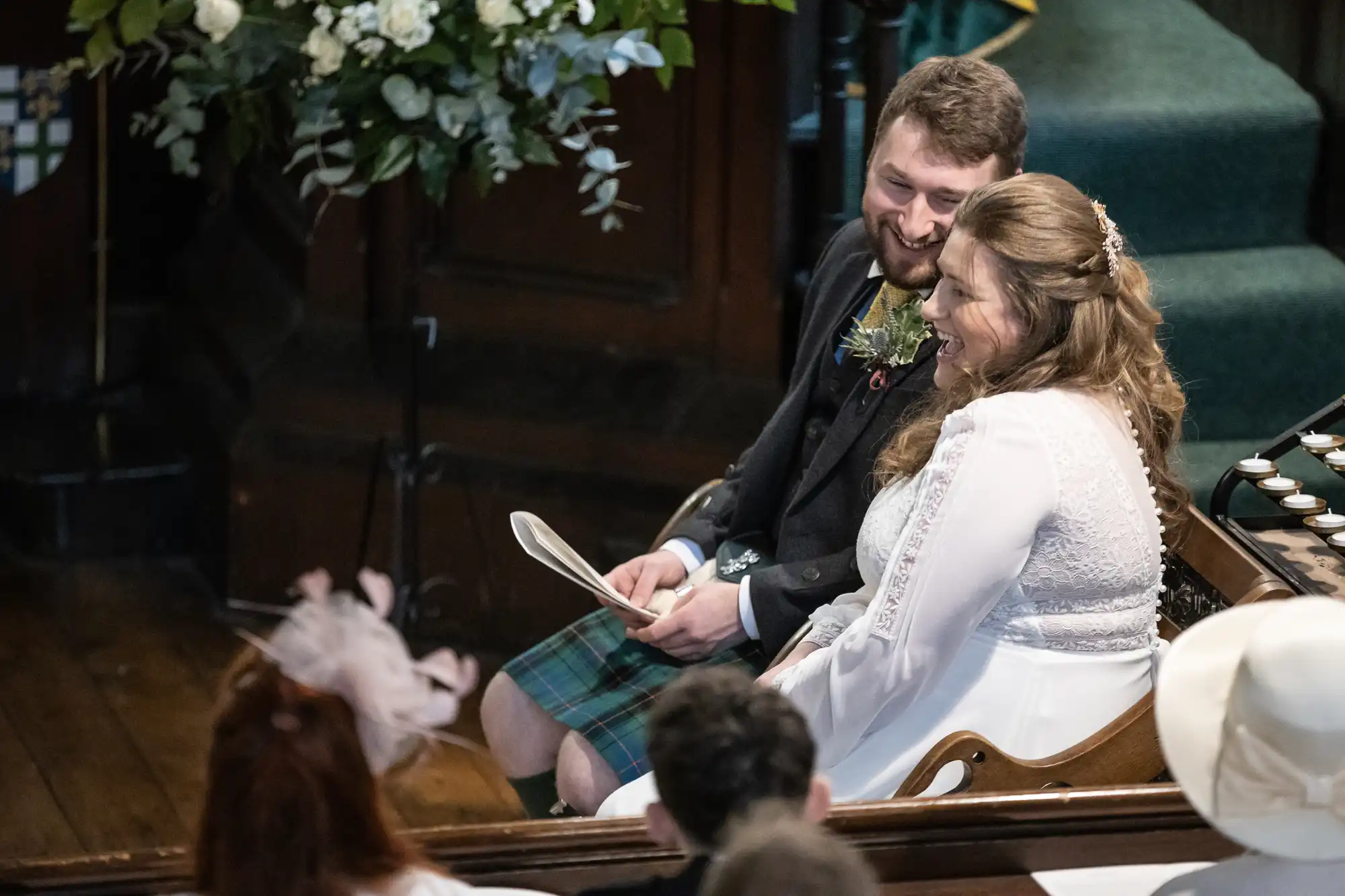 A smiling couple sits together during a wedding ceremony, with the man wearing a kilt and holding a paper, and the woman in a white dress. They are surrounded by family and friends.