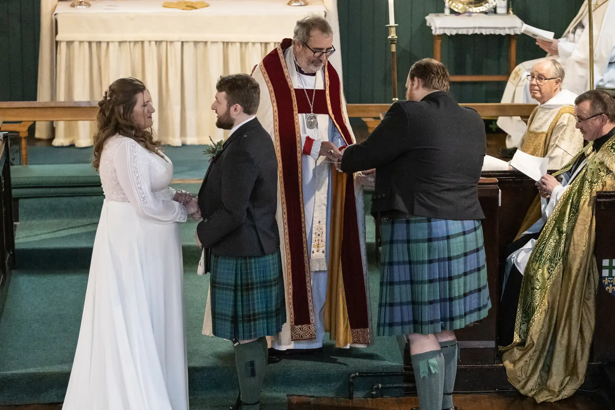 A bride and groom stand at the altar during a wedding ceremony. The officiant and another man, both in traditional attire, are also present at the front of the church.