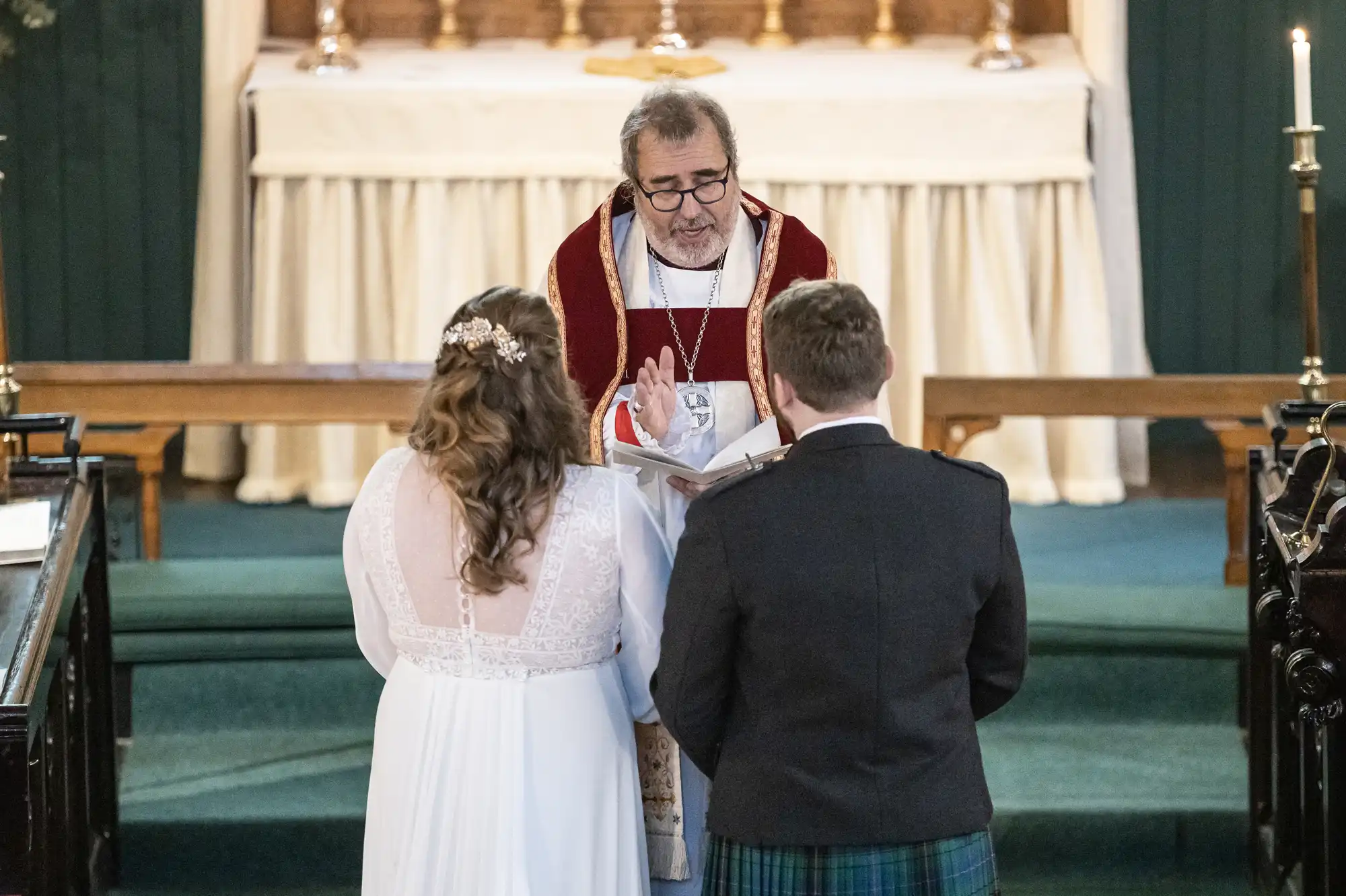 A cleric in religious attire officiates a wedding ceremony, with a bride in a white dress and a groom in a dark suit facing him at the altar.