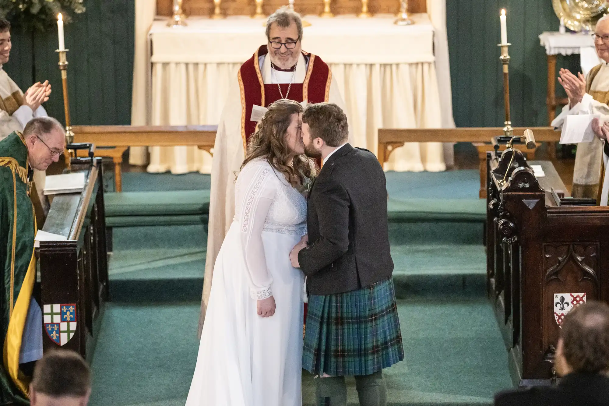 A bride and groom share a kiss at the altar during their wedding ceremony, while a priest and attendants look on in a church setting.