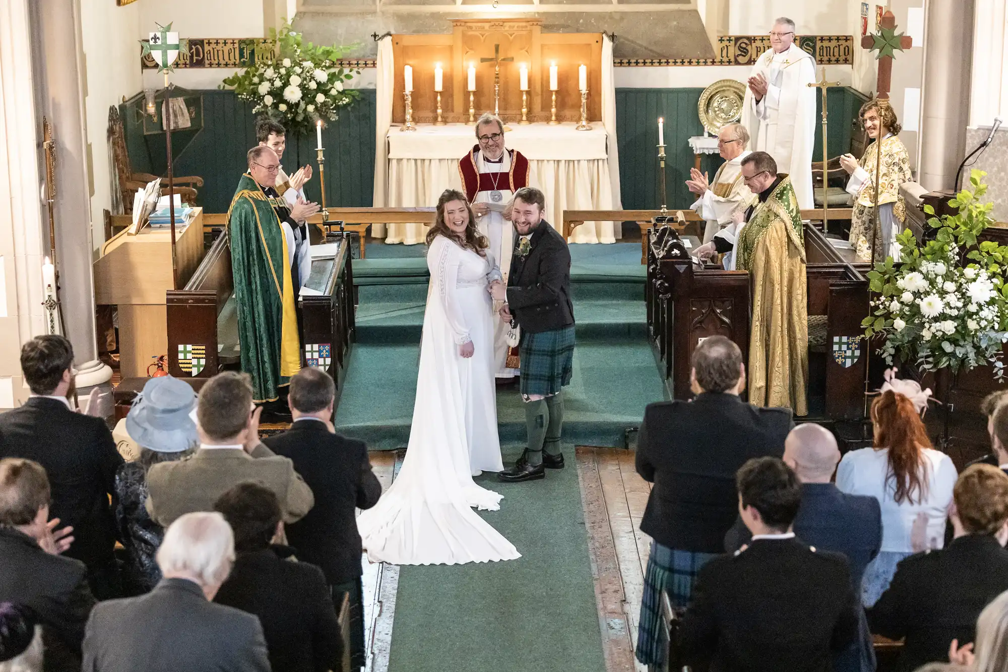 A bride and groom stand at the altar in a church, surrounded by clergy and guests, during a wedding ceremony. The bride wears a white dress, and the groom wears traditional Scottish attire.