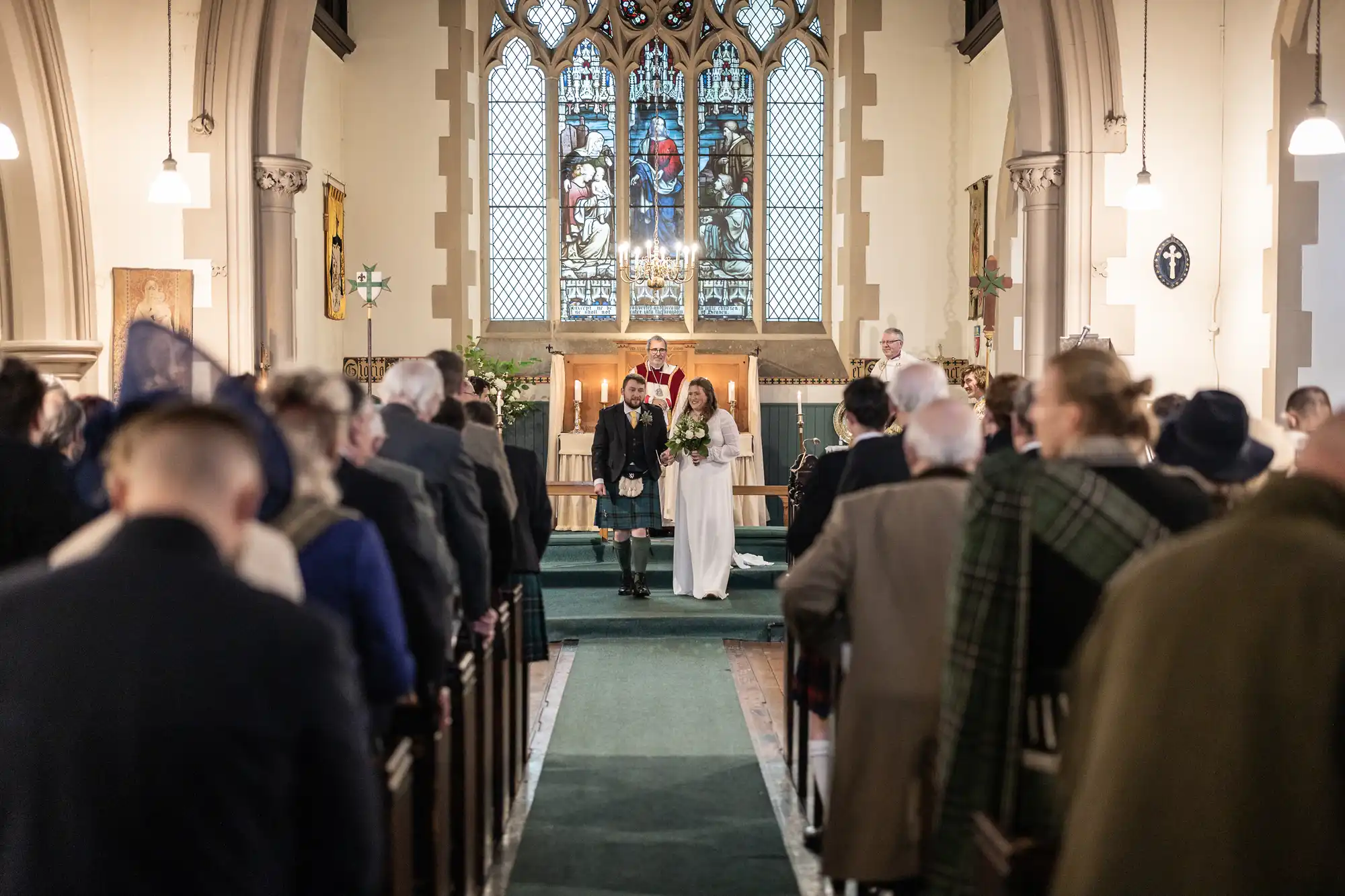 A bride and groom walk down the aisle of a church during a wedding ceremony. The groom is dressed in traditional Scottish attire, and guests stand on either side.