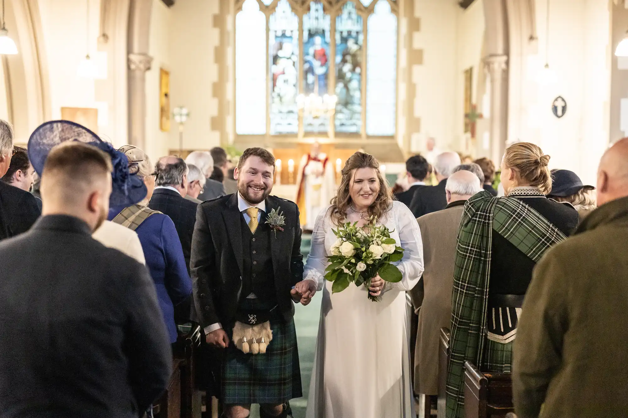 A smiling bride and groom walk down an aisle in a church, holding hands and a bouquet, while guests look on. Scottish kilts and formal attire are worn by some attendees.