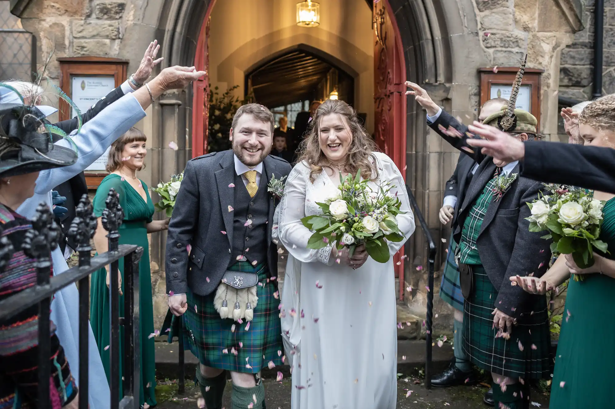 A newlywed couple exits a stone church as guests toss flower petals. Both wear traditional Scottish attire; the bride holds a bouquet while the groom wears a kilt. Guests line the path, celebrating.