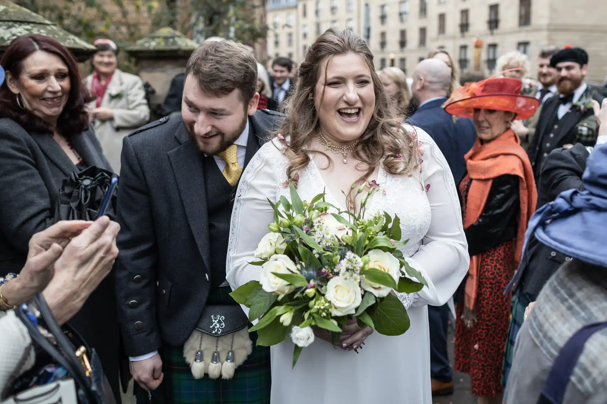 A bride in a white dress holding a bouquet stands smiling next to a man in a traditional kilt amidst a crowd of people.