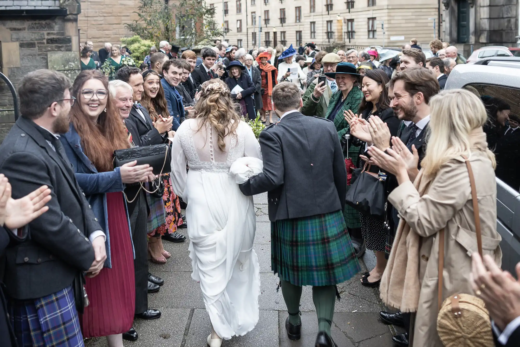 A newlywed couple in traditional Scottish attire walks between cheering guests outdoors, with the bride wearing a white gown and the groom in a kilt.