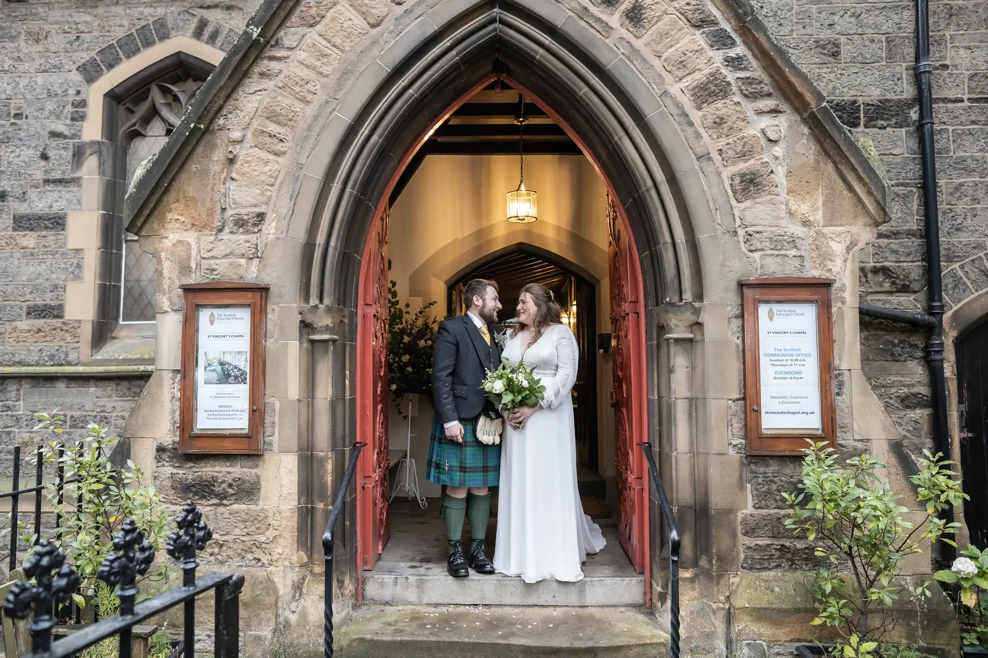 A couple in wedding attire stands at the entrance of a stone church, looking at each other. The groom wears a kilt, while the bride holds a bouquet of flowers.