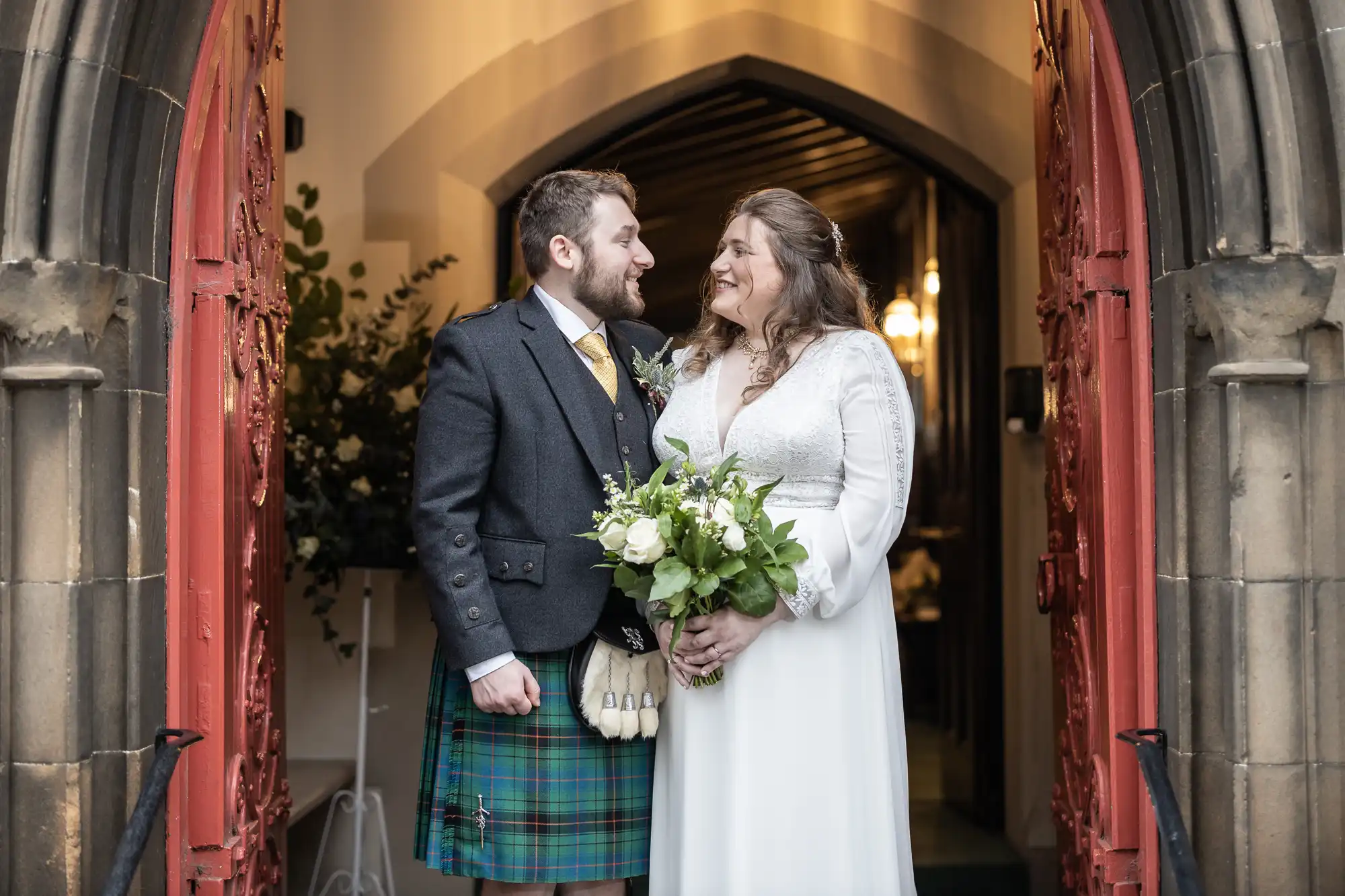 A couple, the groom in a kilt and the bride in a white dress, stand smiling at each other in a church doorway, holding a bouquet of flowers.