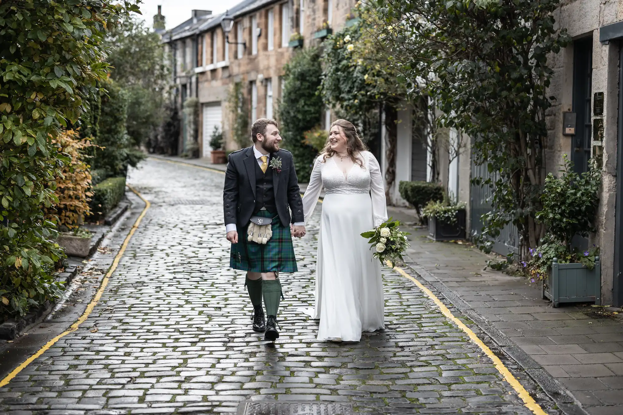 A couple in wedding attire, with the groom in a kilt and the bride in a white dress, walk hand-in-hand on a cobblestone street lined with greenery.