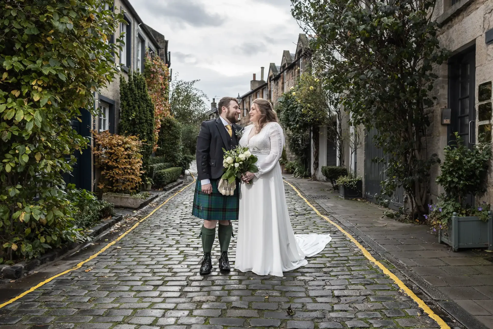 St Vincent's Chapel wedding photographer image of a bride in a white dress and a groom in a kilt hold hands and stand on a cobblestone street lined with townhouses and greenery.