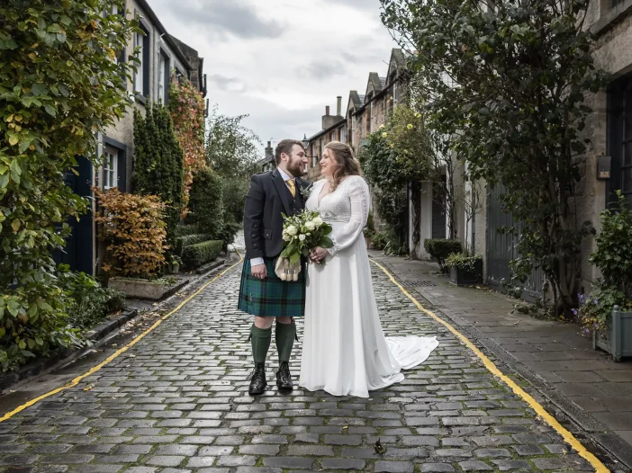 A bride in a white dress and a groom in a kilt hold hands and stand on a cobblestone street lined with townhouses and greenery.