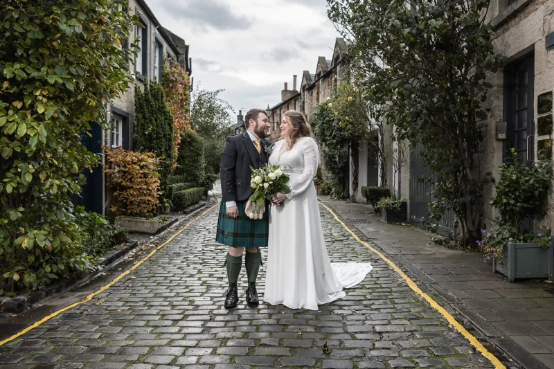 A bride in a white dress and a groom in a kilt hold hands and stand on a cobblestone street lined with townhouses and greenery.