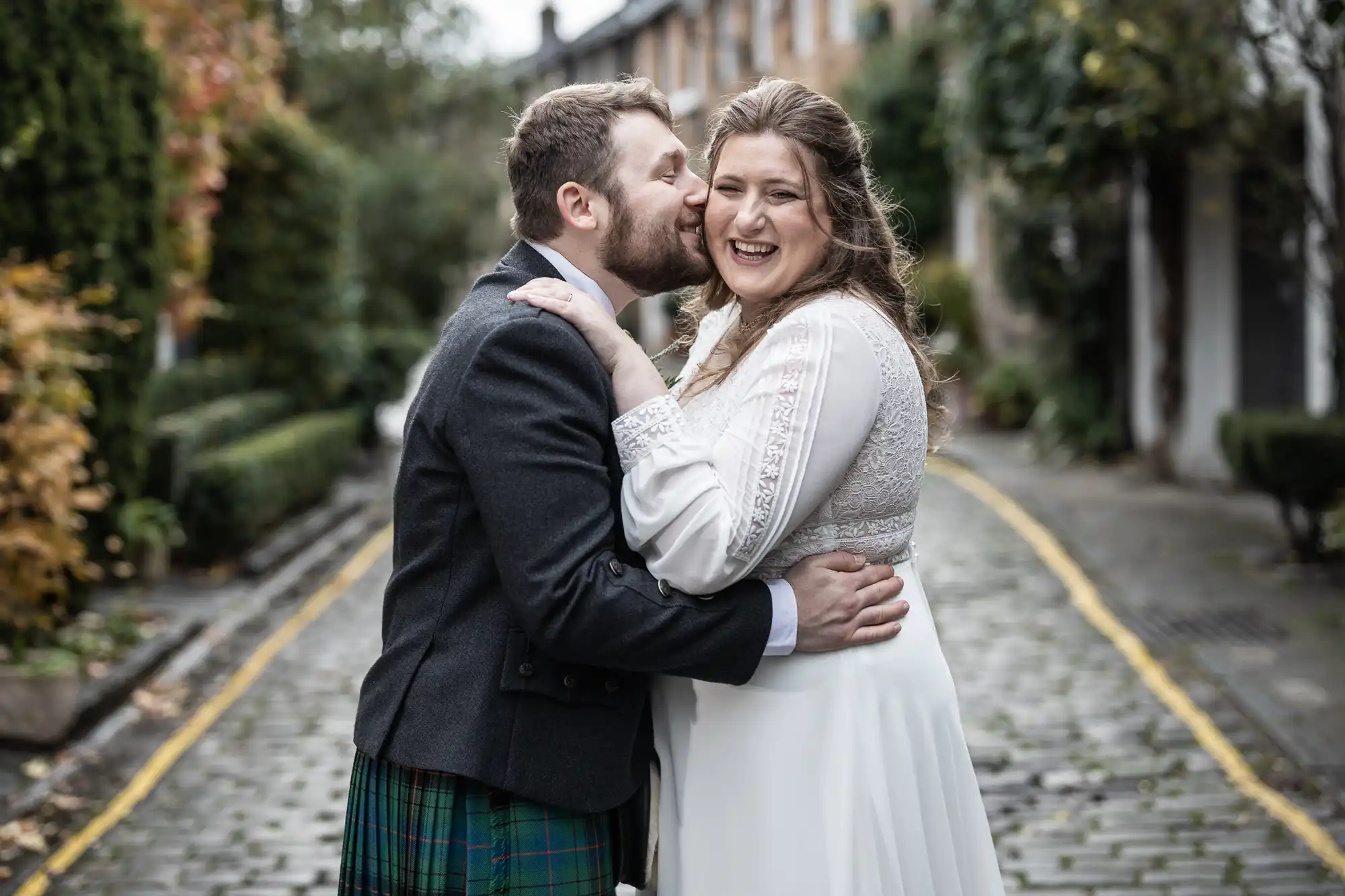 A man kisses a smiling woman on the cheek as they embrace on a cobblestone street lined with greenery and houses.