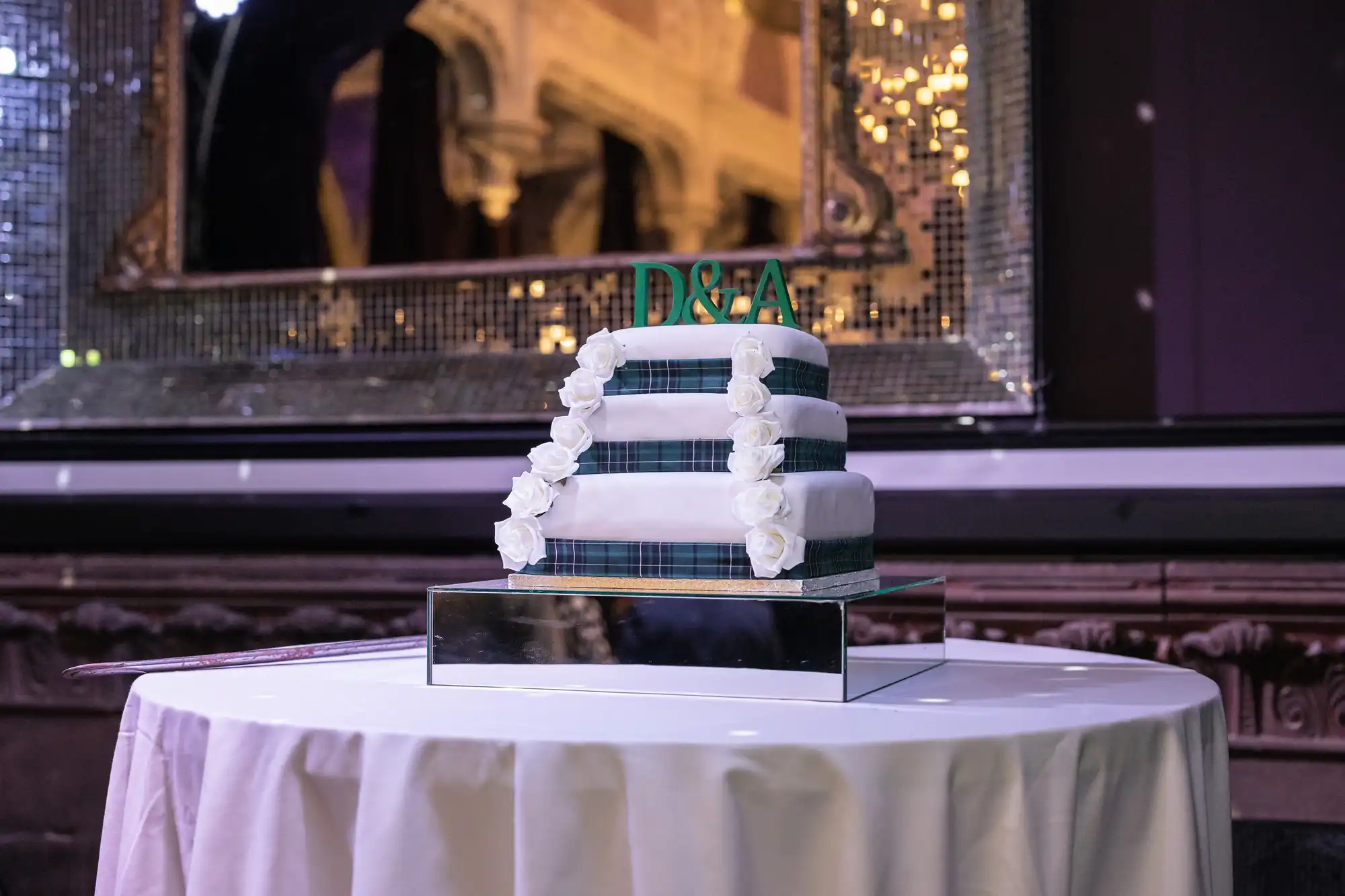 A three-tiered cake decorated with white flowers and green ribbon sits on a round table, with "D&A" initials on top. The cake is placed in front of a large, ornate mirror with a mosaic frame.