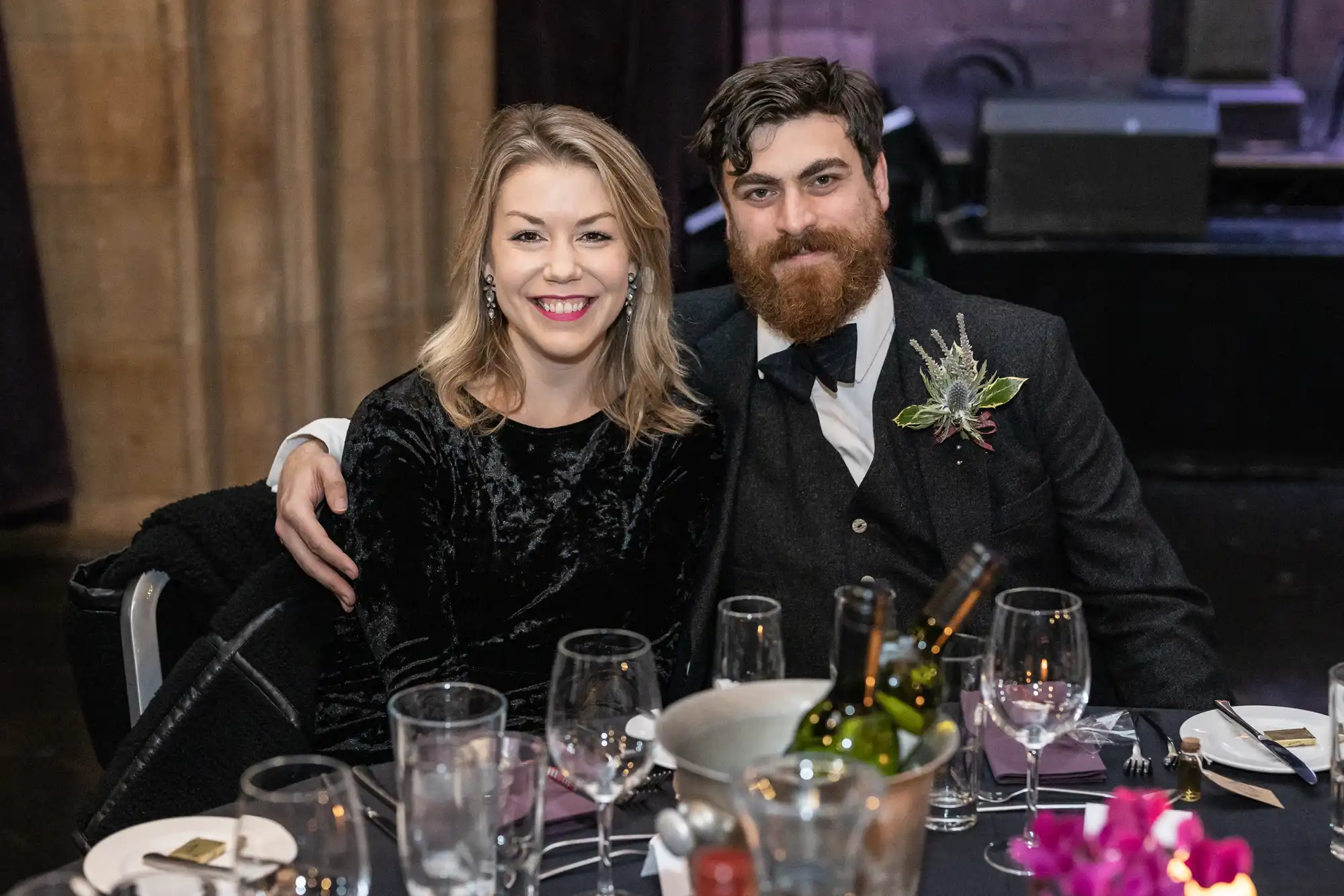 Ghillie Dhu wedding reception - A woman and a man, dressed formally, sit together at a table with empty wine glasses and a bucket with wine bottles. The man has his arm around the woman, and both are smiling at the camera.